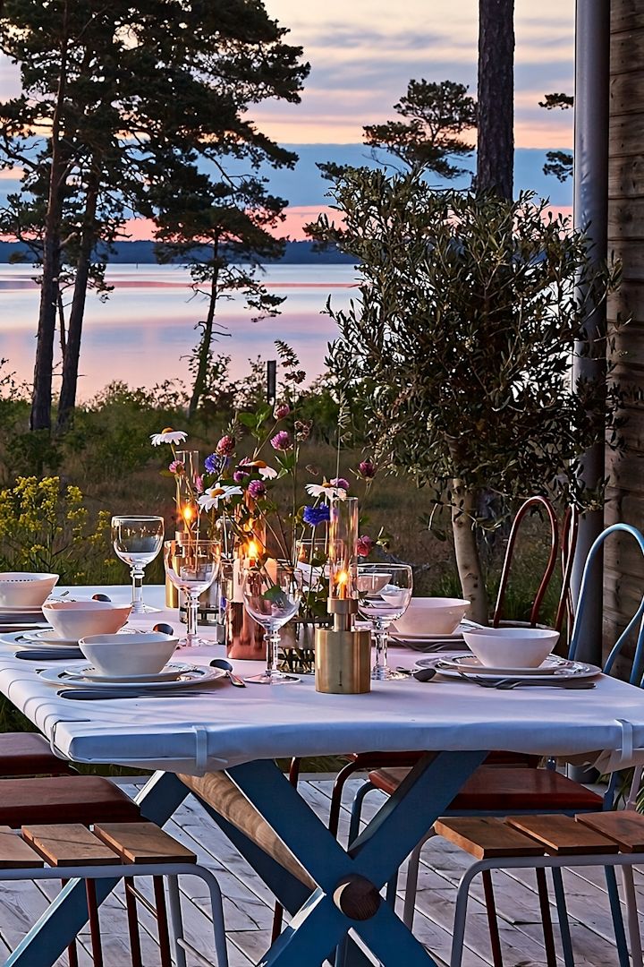 Lanternes et photophores pour l'extérieur - ici, vous voyez une table en bois sur une terrasse avec la mer en arrière-plan pendant le coucher du soleil. La table est dressée et décorée, entre autres, avec la mini-lampe à huile Patina de Klong.