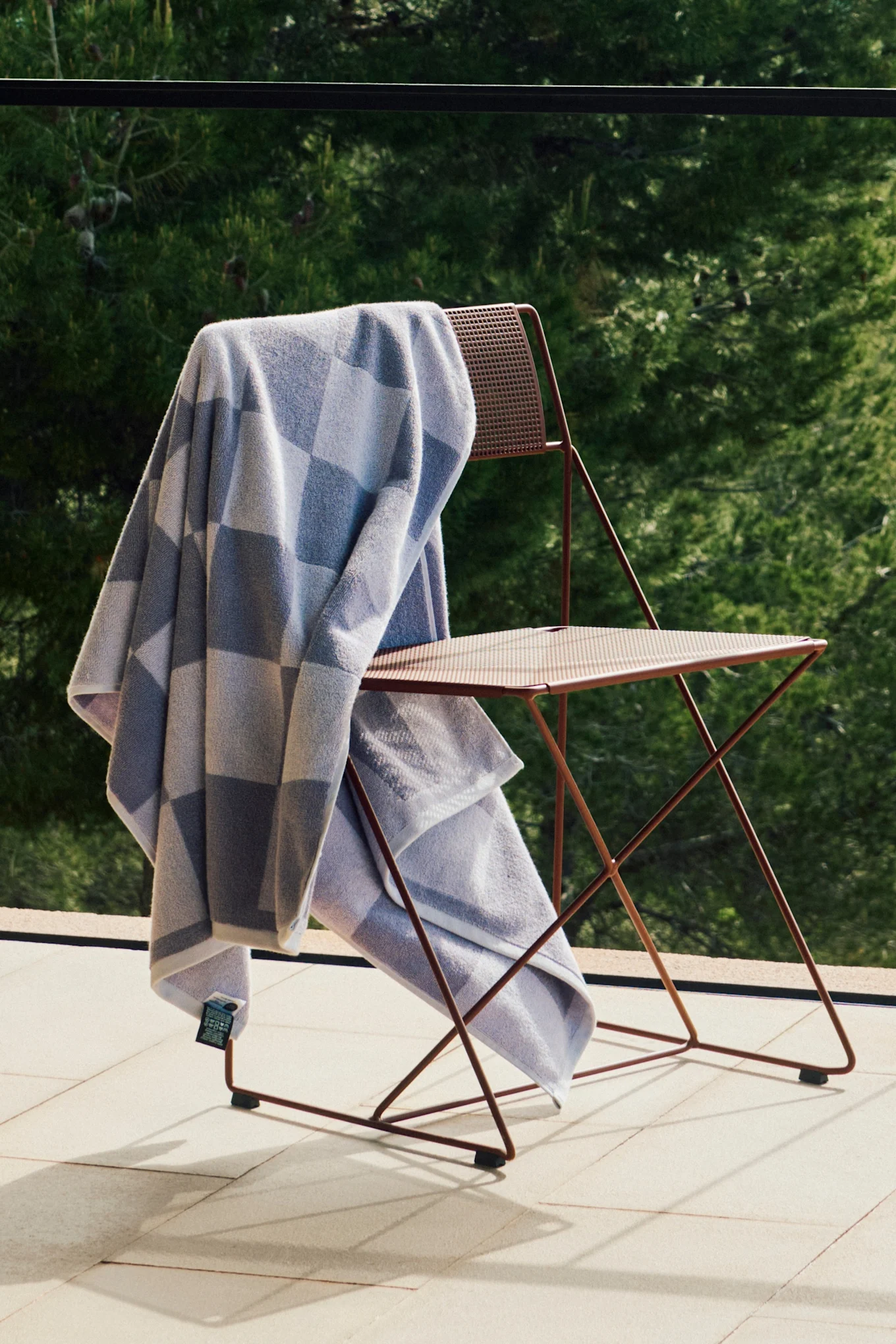 Serviette à carreaux bleu clair et gris sur une chaise pliante en métal brun rougeâtre sur une terrasse ensoleillée avec de la verdure.