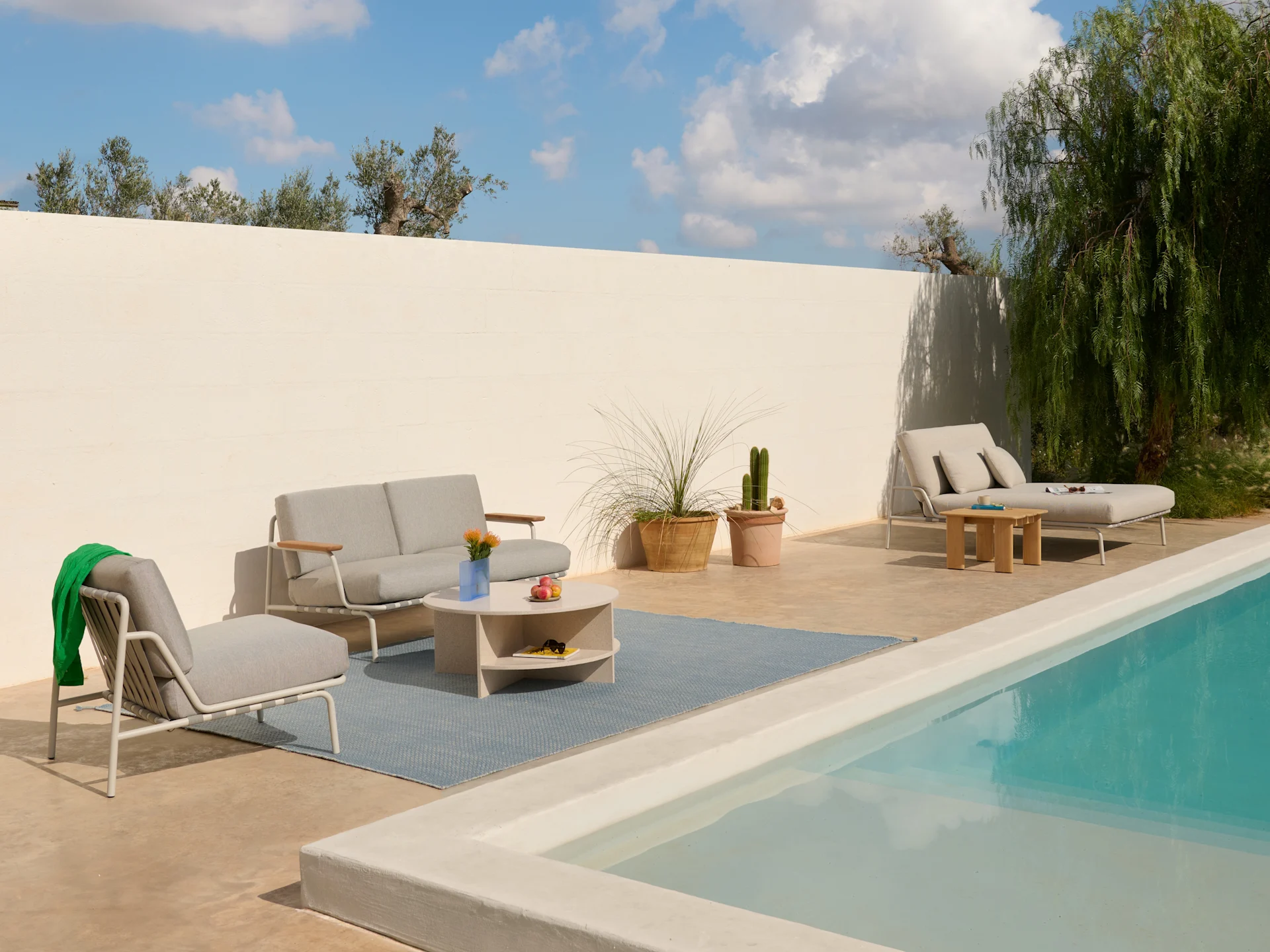 Terrasse extérieure avec canapé gris clair, fauteuils, table basse sur tapis bleu et chaise longue au bord d'une piscine.