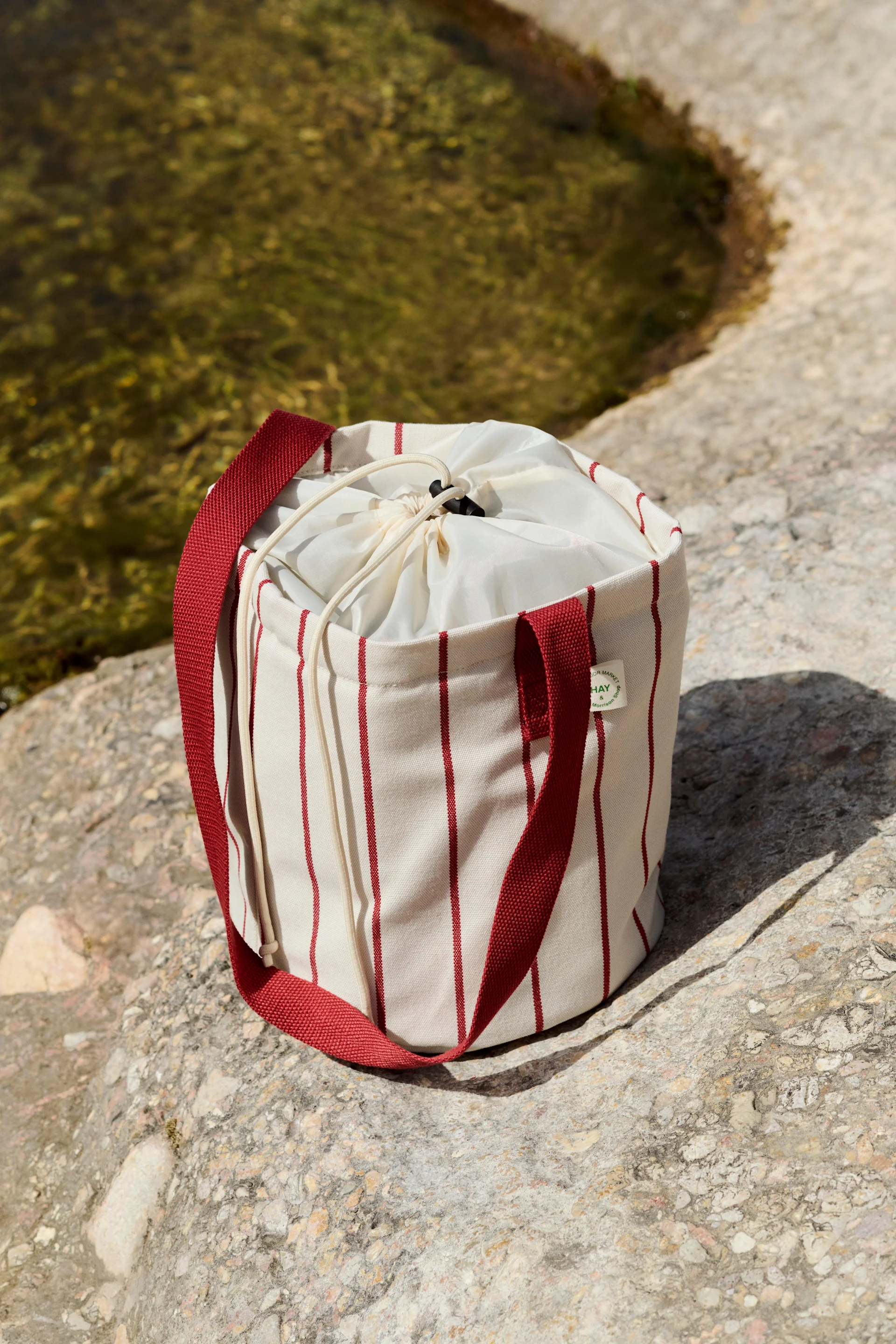Sac cylindrique crème à rayures verticales rouges et fermeture à cordon, posé sur des rochers au bord de l'eau.