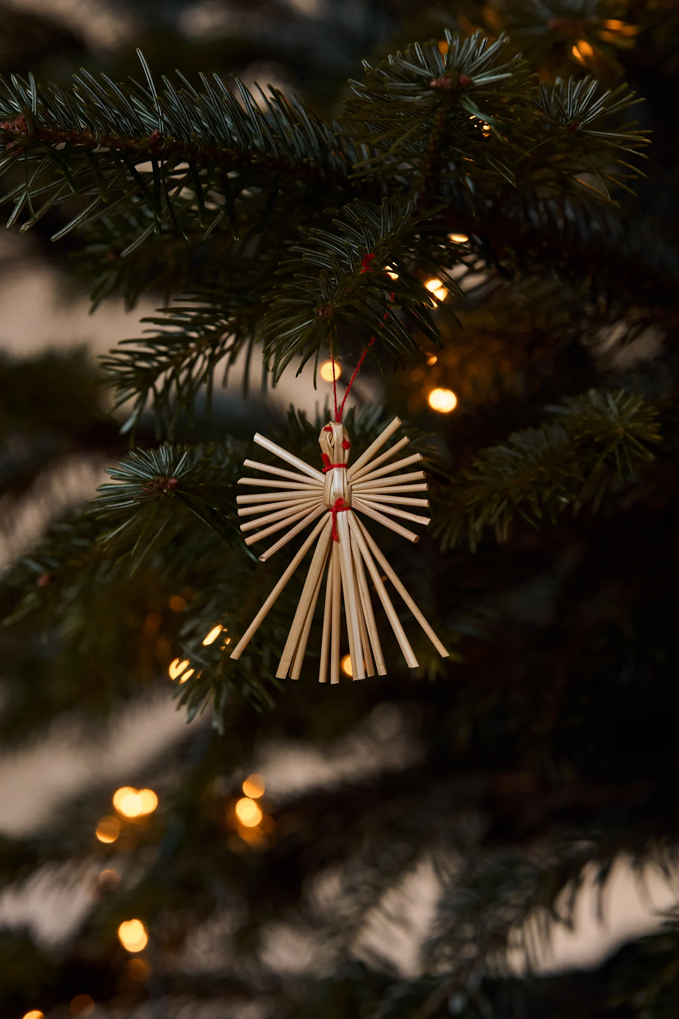Ornement d'ange en paille suspendu à une branche de sapin de Noël avec des lumières floues en arrière-plan.