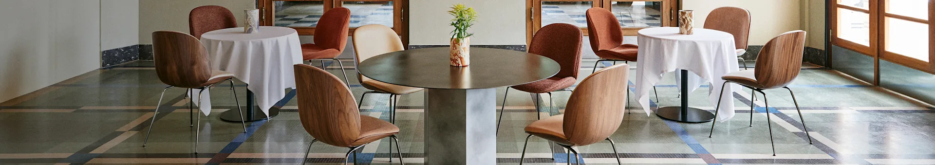 Intérieur de café élégant avec une grande table centrale, deux petites tables avec des nappes blanches et diverses chaises sur un sol à motifs.