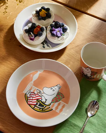 Assiette, tasse et cuillère Moomintroll à côté d'une assiette de meringues aux myrtilles et fleurs sur une table en bois.