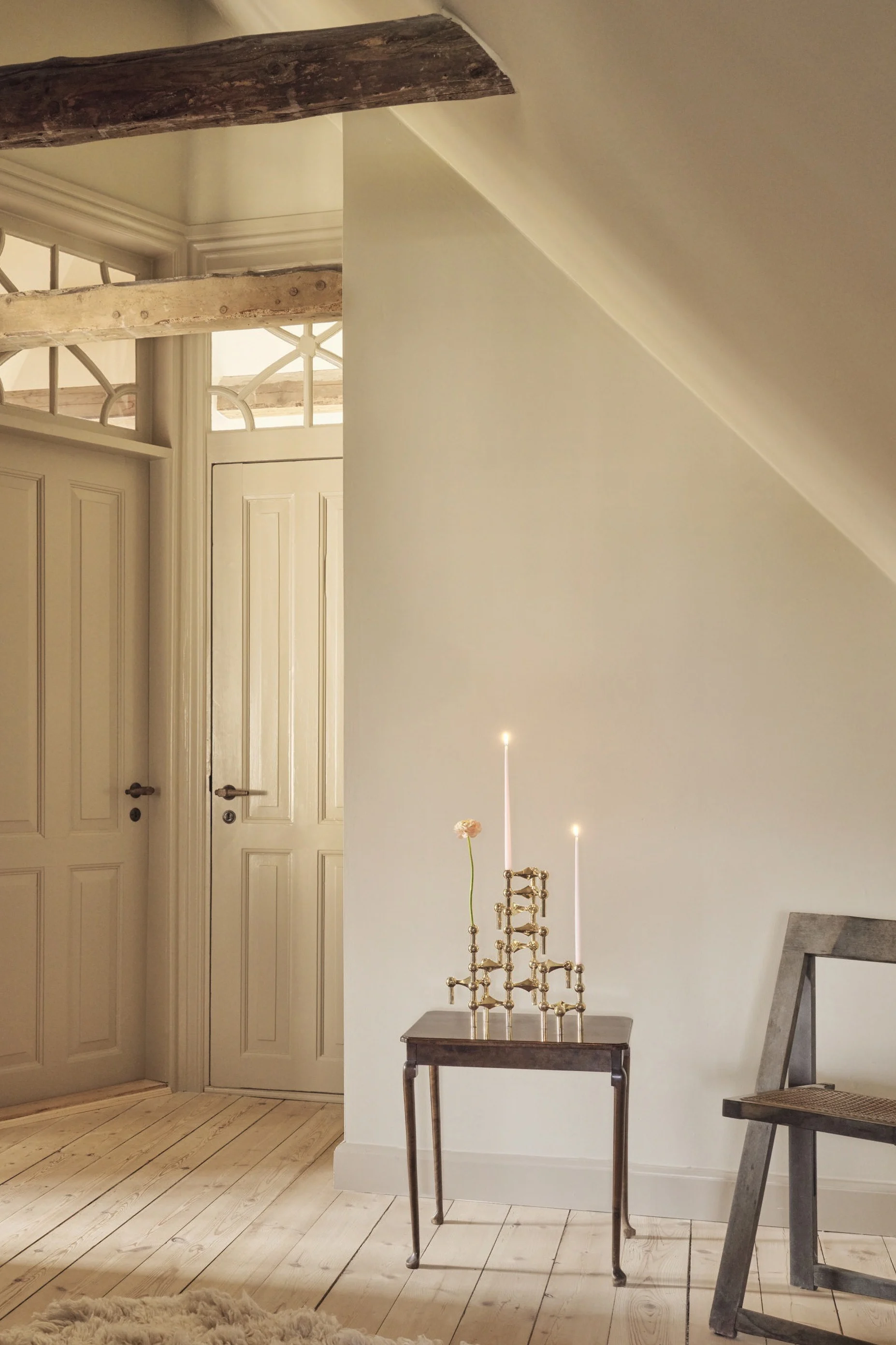 Une pièce mansardée avec des murs clairs et des planchers en bois. Deux portes à panneaux avec impostes ornées, une poutre apparente, une petite table d'appoint avec des bougies allumées et une fleur, une chaise et un tapis moelleux.