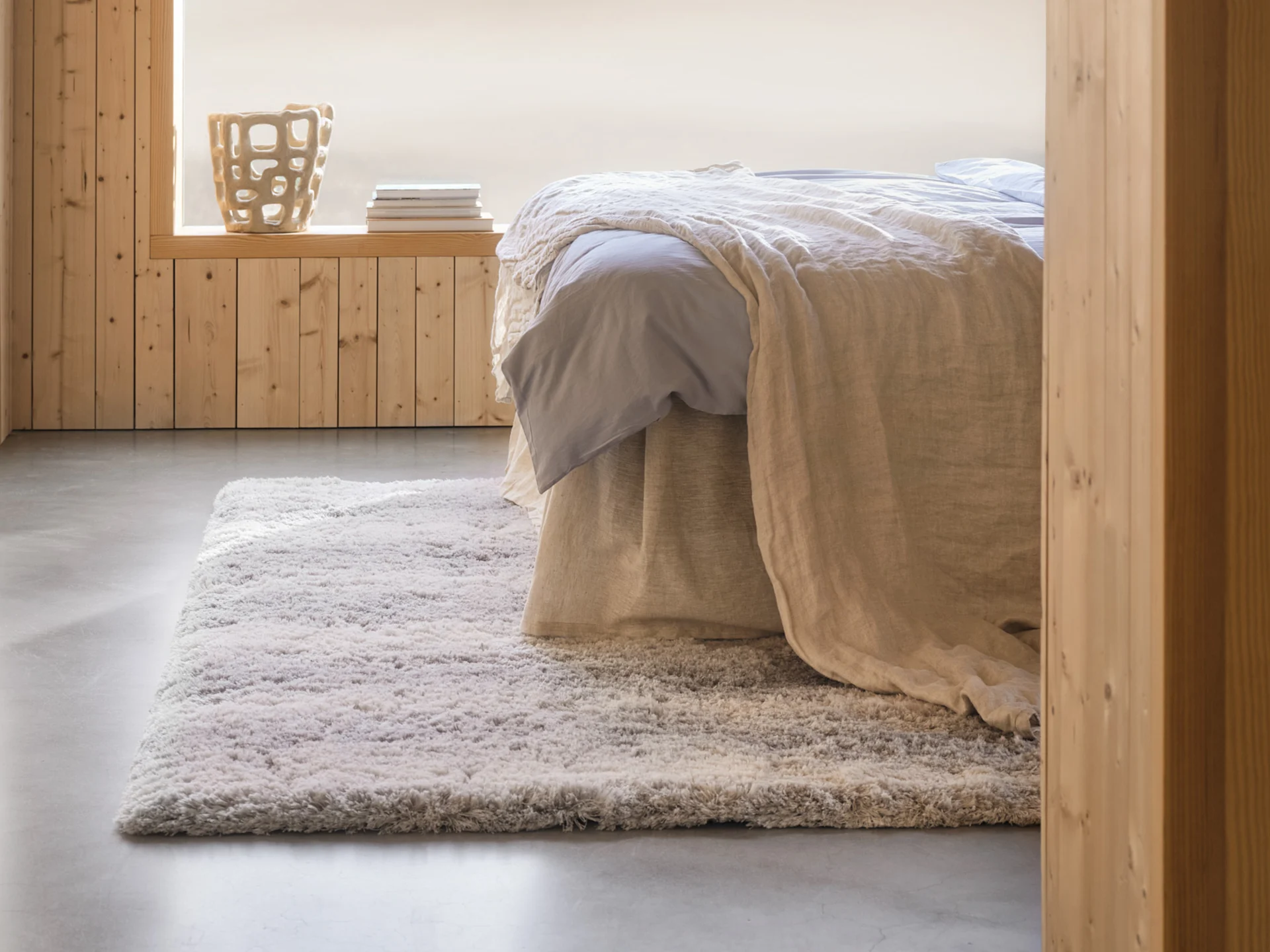 Chambre douillette avec un lit, des draps bleus clairs, une couverture beige, un tapis shaggy, des murs en bois et un vase décoratif.