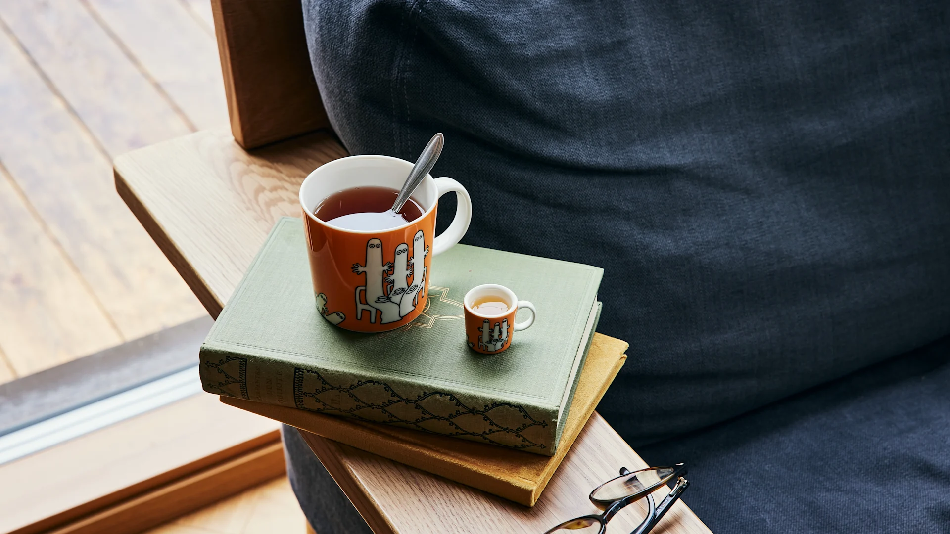 Le mug Hatifnatten du personnage Moomin, de couleur orange, avec du thé, se trouve à côté d'un canapé, sur une pile de livres.