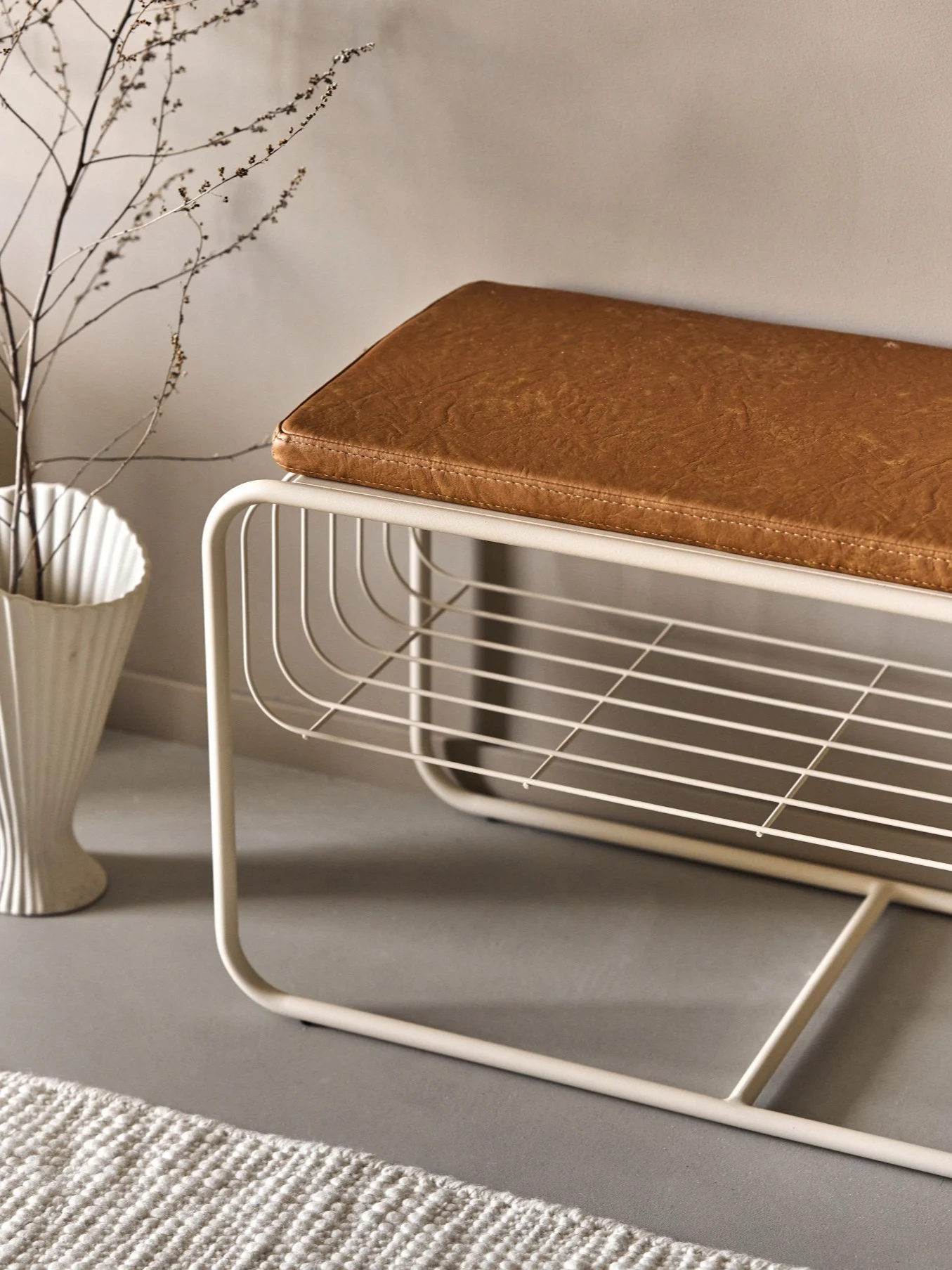 Banc moderne en métal beige avec coussin en similicuir marron, doté d'une étagère à lattes ouverte. Un vase blanc avec des branches et un tapis sont à proximité.
