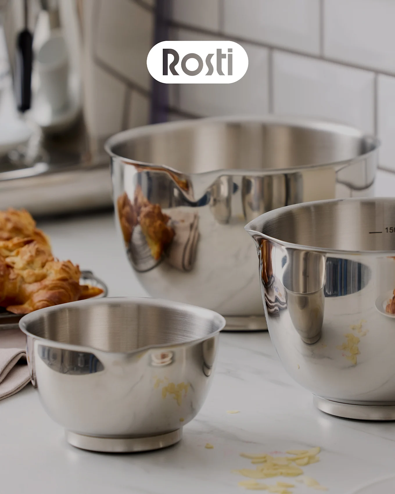Trois bols Rosti sur un comptoir : un bol beige avec un bol bleu empilé dessus, les deux avec des couvercles, et un petit bol jaune rempli d'abricots secs.