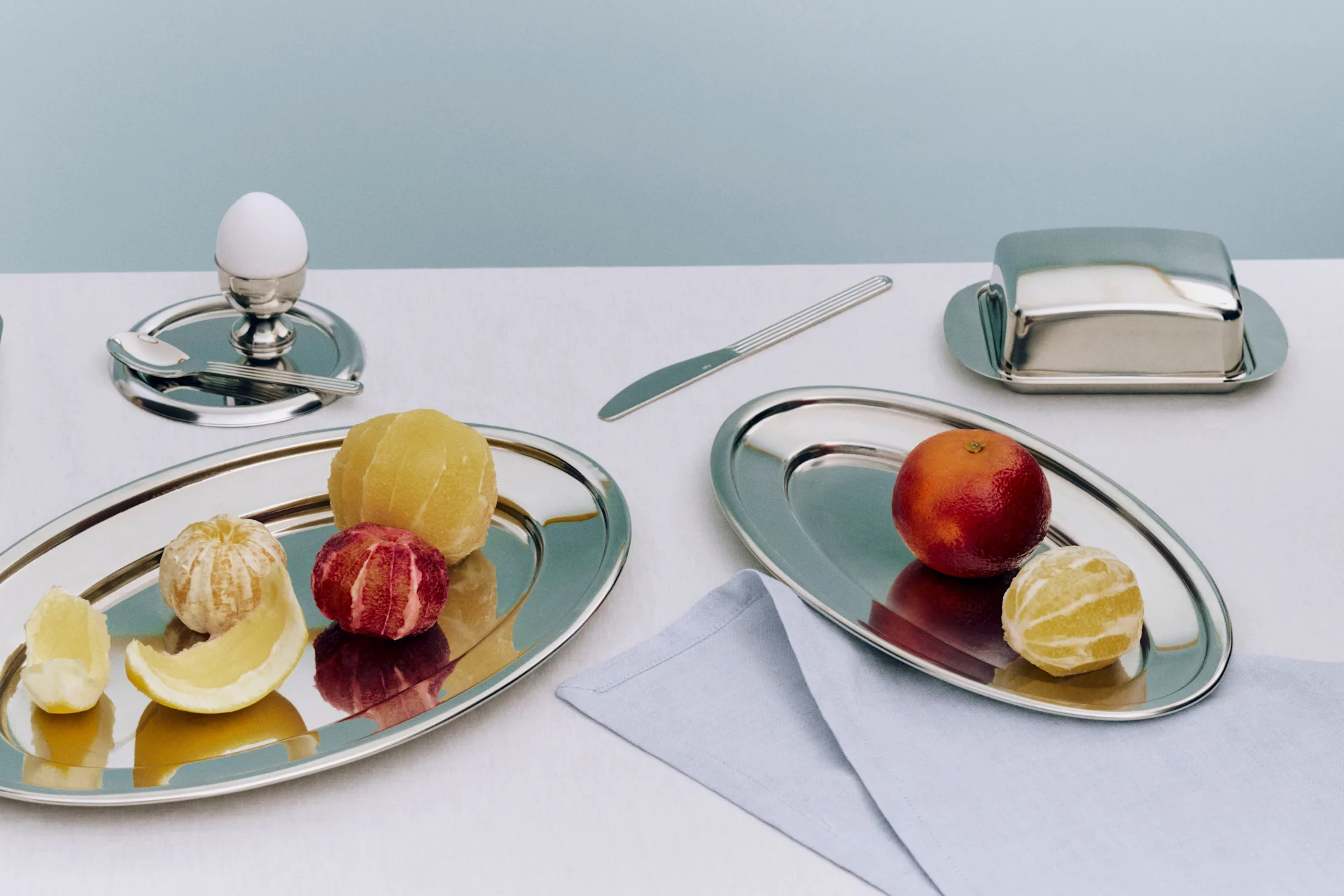 Un petit-déjeuner avec deux plateaux argentés contenant des agrumes pelés, un œuf dans un coquetier, un beurrier et un couteau.