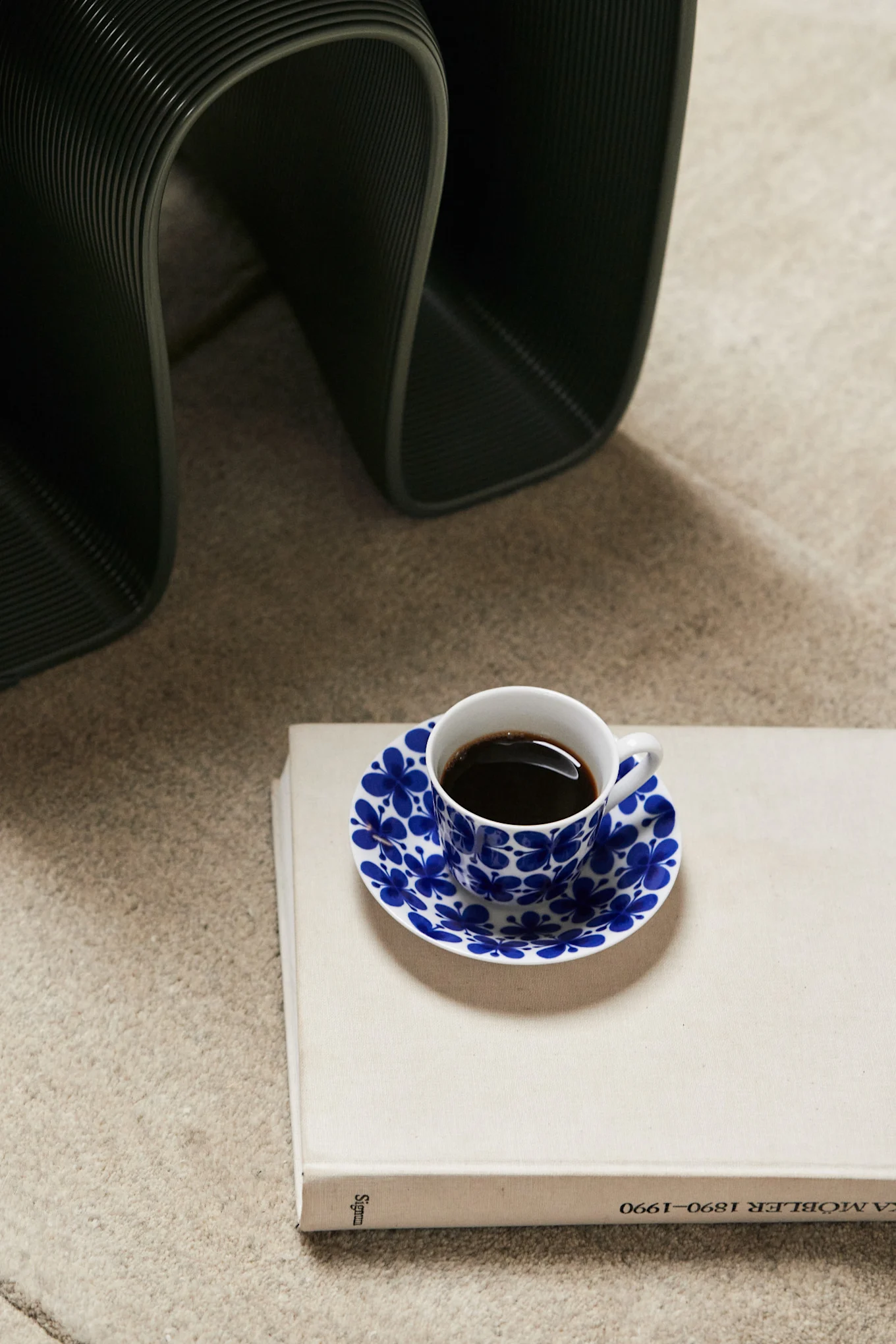 Tasse à café avec soucoupe florale bleue sur un livre clair, objet nervuré foncé en arrière-plan.