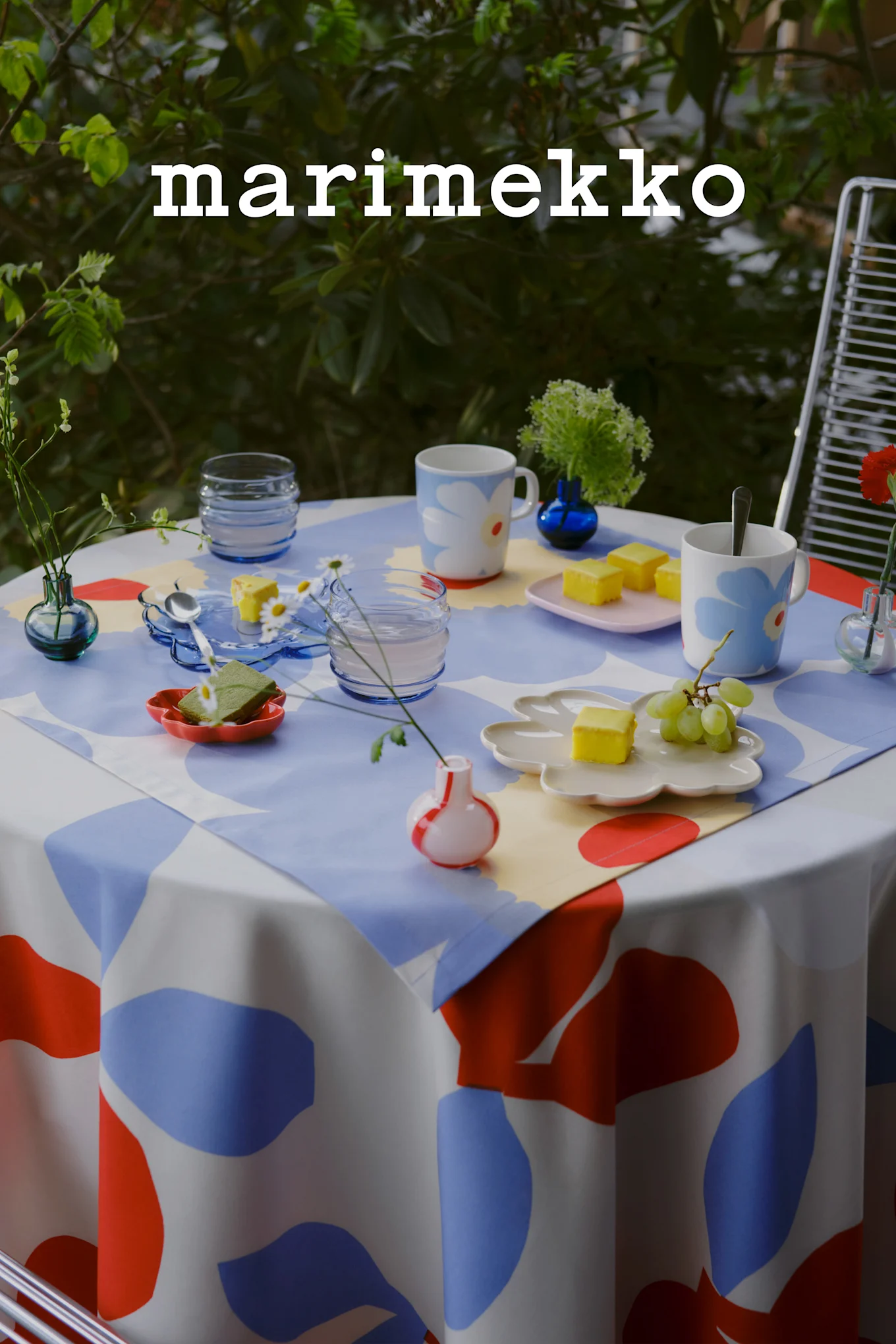 Logo Marimekko. Une table d'extérieur dressée avec une nappe à motifs abstraits bleus, rouges et blancs, des mugs floraux, des gâteaux jaunes, des raisins et de petits vases.
