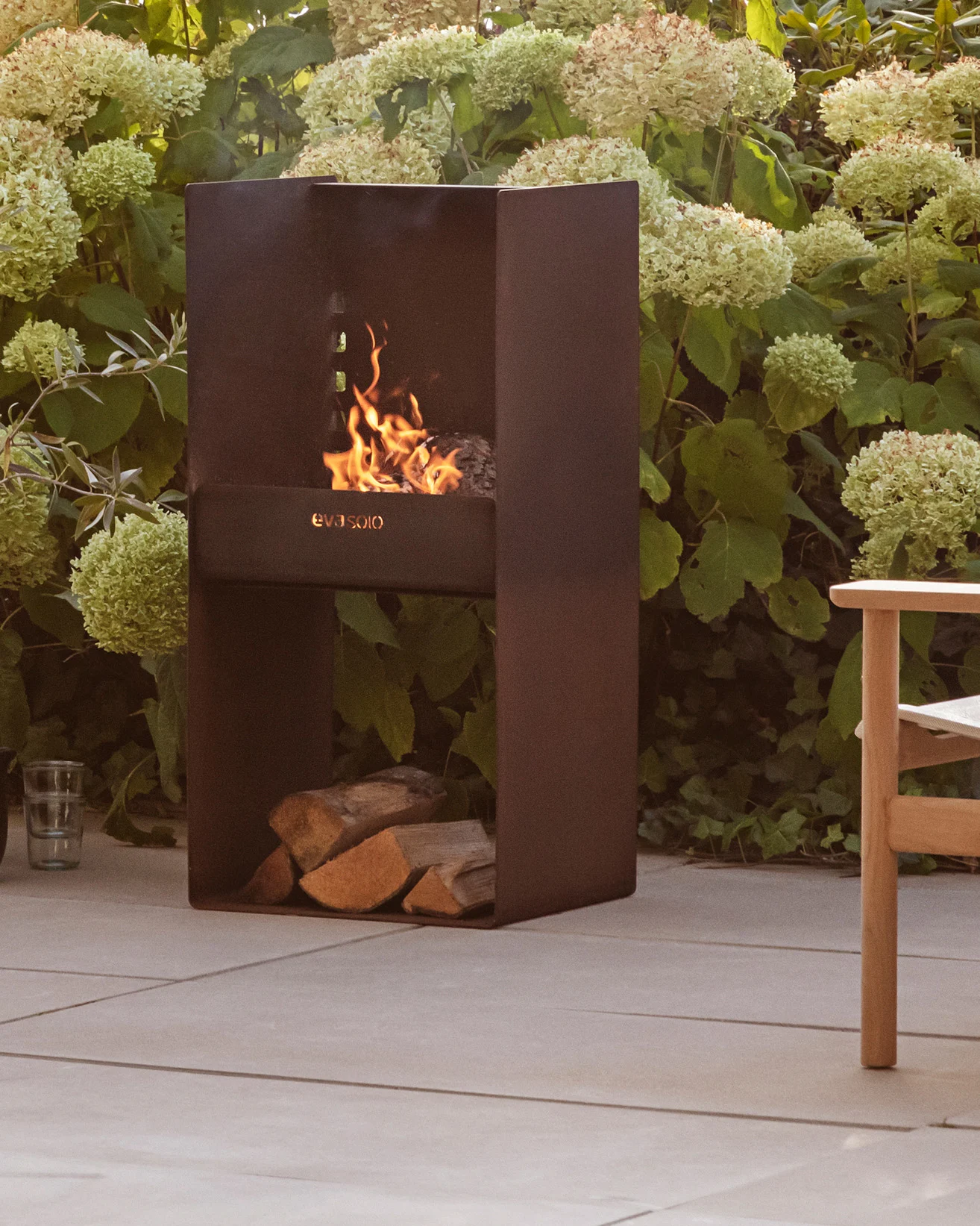 Cheminée d'extérieur moderne en métal foncé avec un feu allumé, bûches empilées en dessous, entourée de plantes vertes et d'un patio.