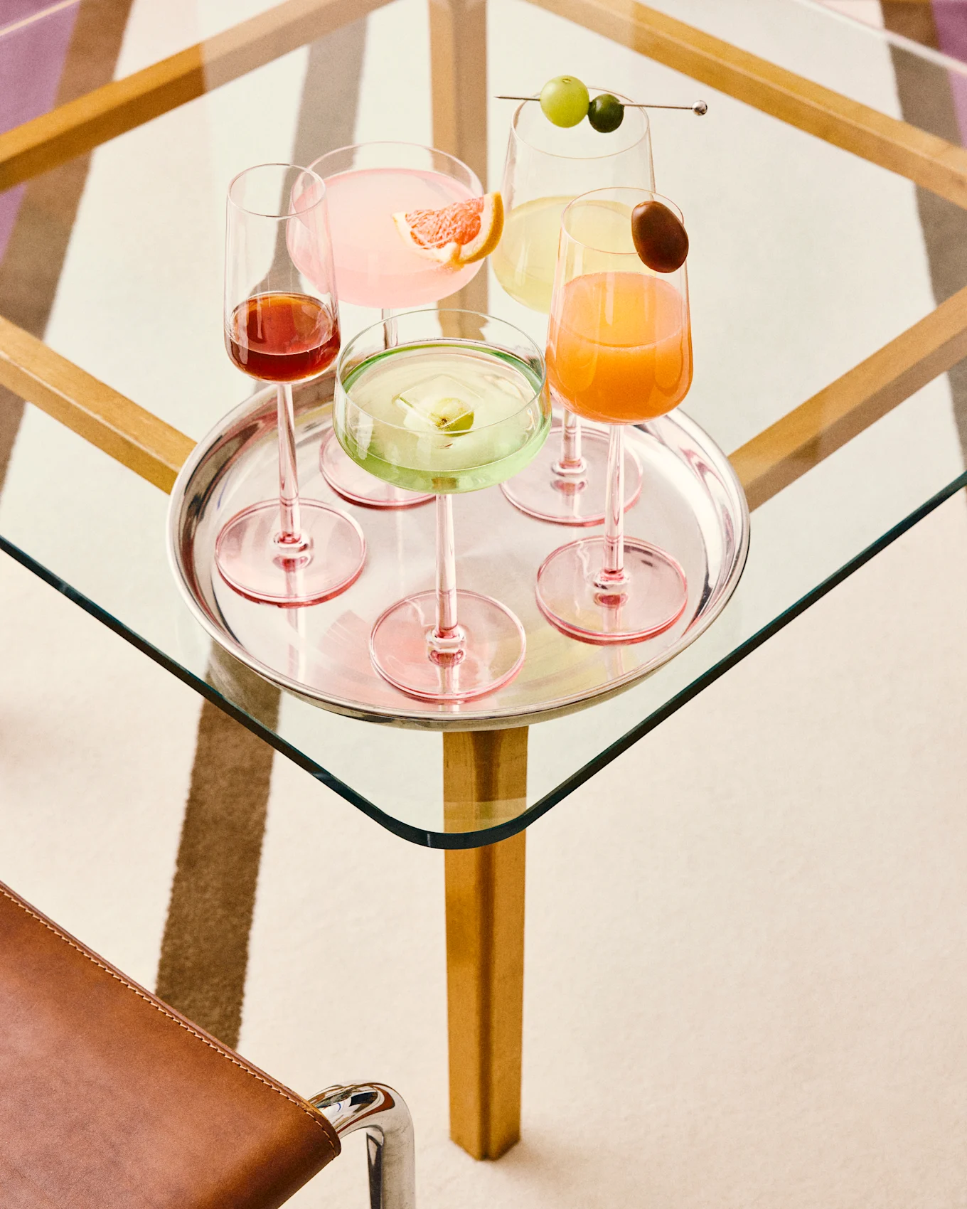 Cinq cocktails colorés sur un plateau en argent : rose au pamplemousse, vert à la glace aux fruits, jaune aux raisins, orange avec une garniture, et rouge foncé, tous sur une table en verre.