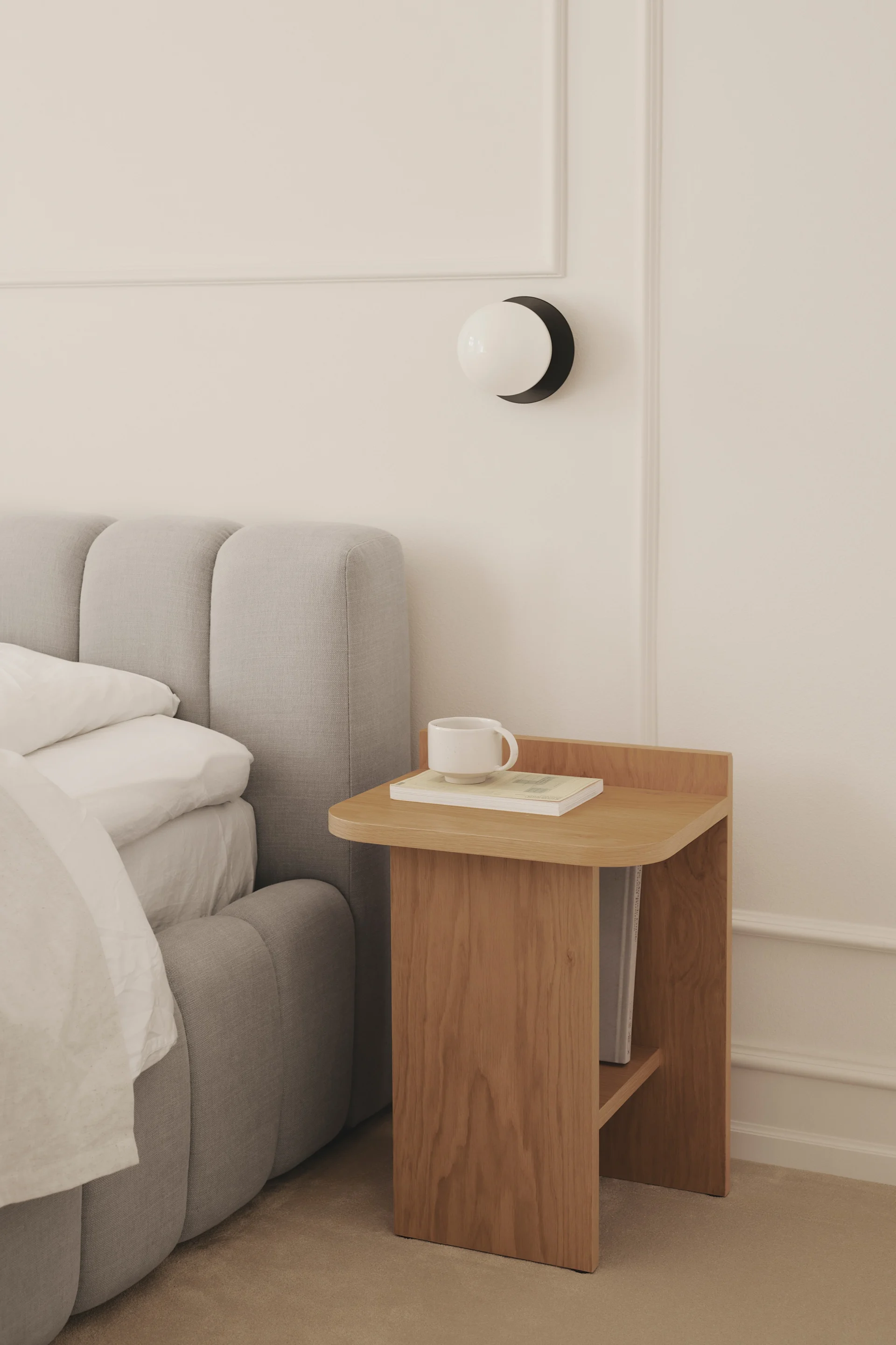 Une chambre douillette avec un lit recouvert d'un linge de lit gris, à côté d'une table de chevet en bois sur laquelle sont posés une tasse et un livre, ainsi qu'une applique murale ronde.