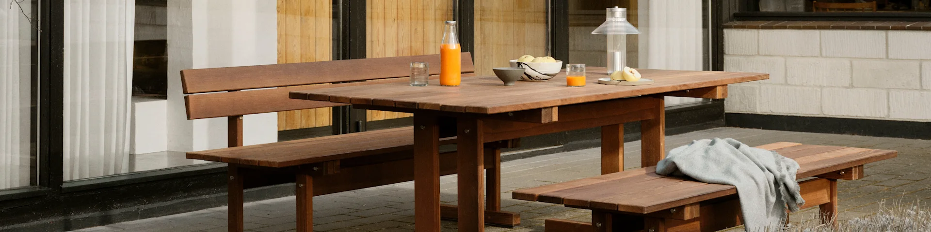 Un ensemble de table de pique-nique en bois avec une bouteille de jus et des fruits, et une couverture grise sur un banc.