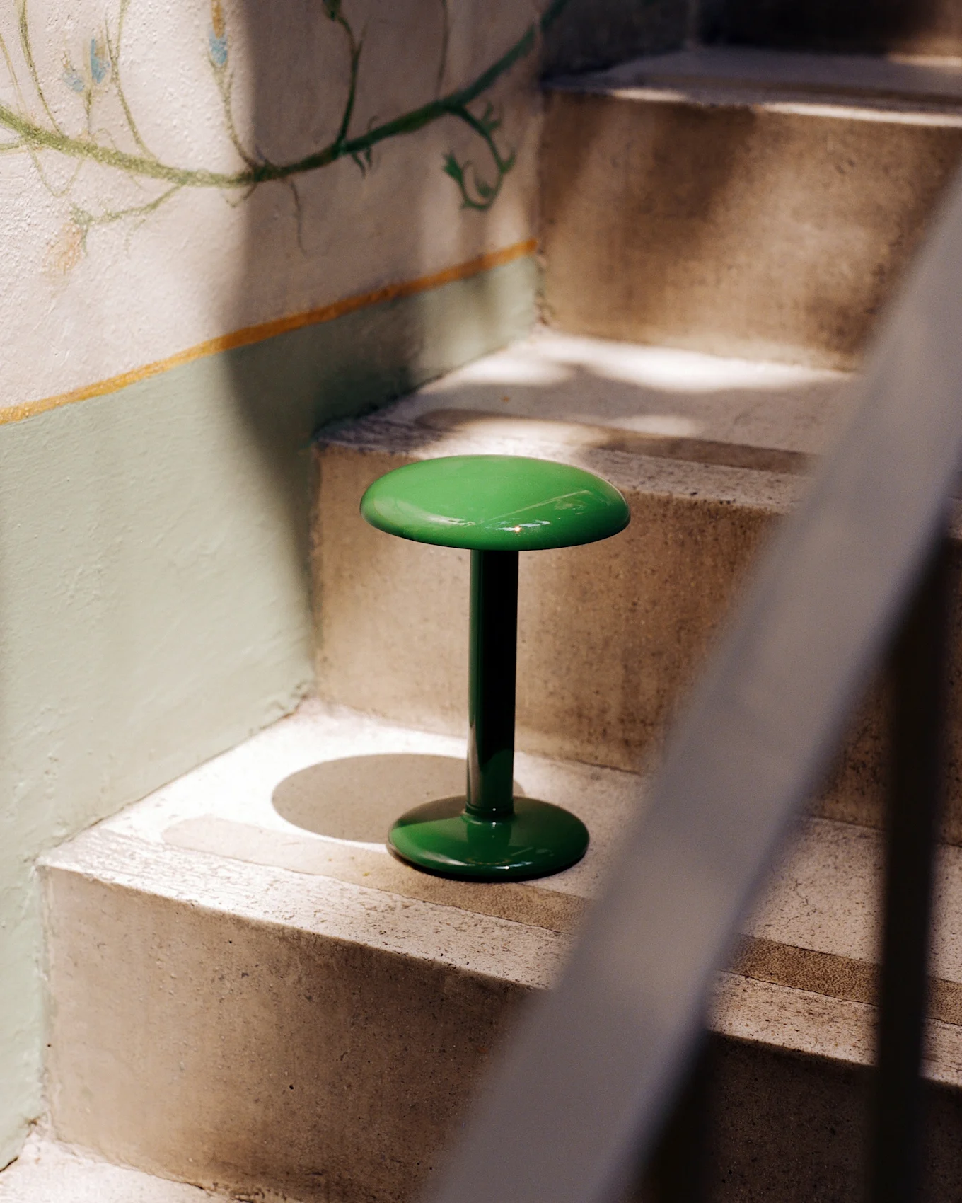 Un tabouret vert vif et brillant avec un plateau et une base ronds se tient sur un escalier en béton à côté d'un mur orné d'une fresque florale fanée.