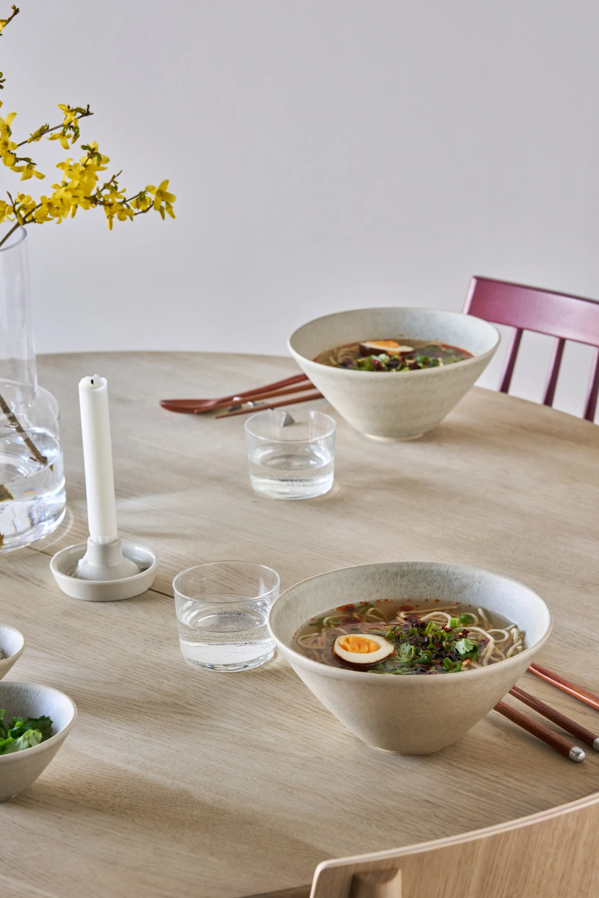 Deux bols de ramen avec œufs et garniture sur une table en bois clair, avec des verres d'eau et des fleurs jaunes dans un vase.