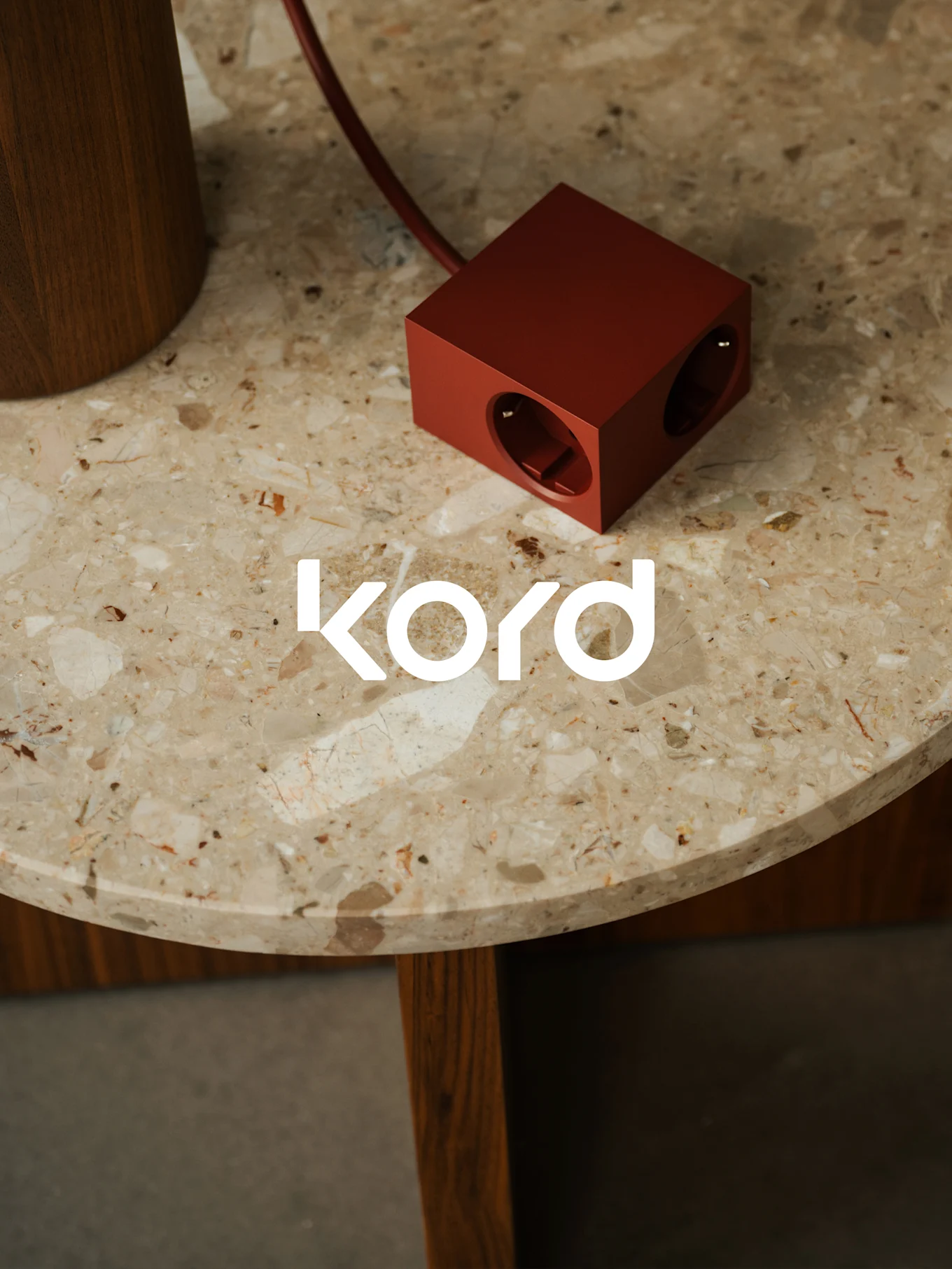 Une multiprise rouge foncé en forme de cube avec deux prises visibles repose sur une table ronde en marbre, avec le logo blanc "kord".