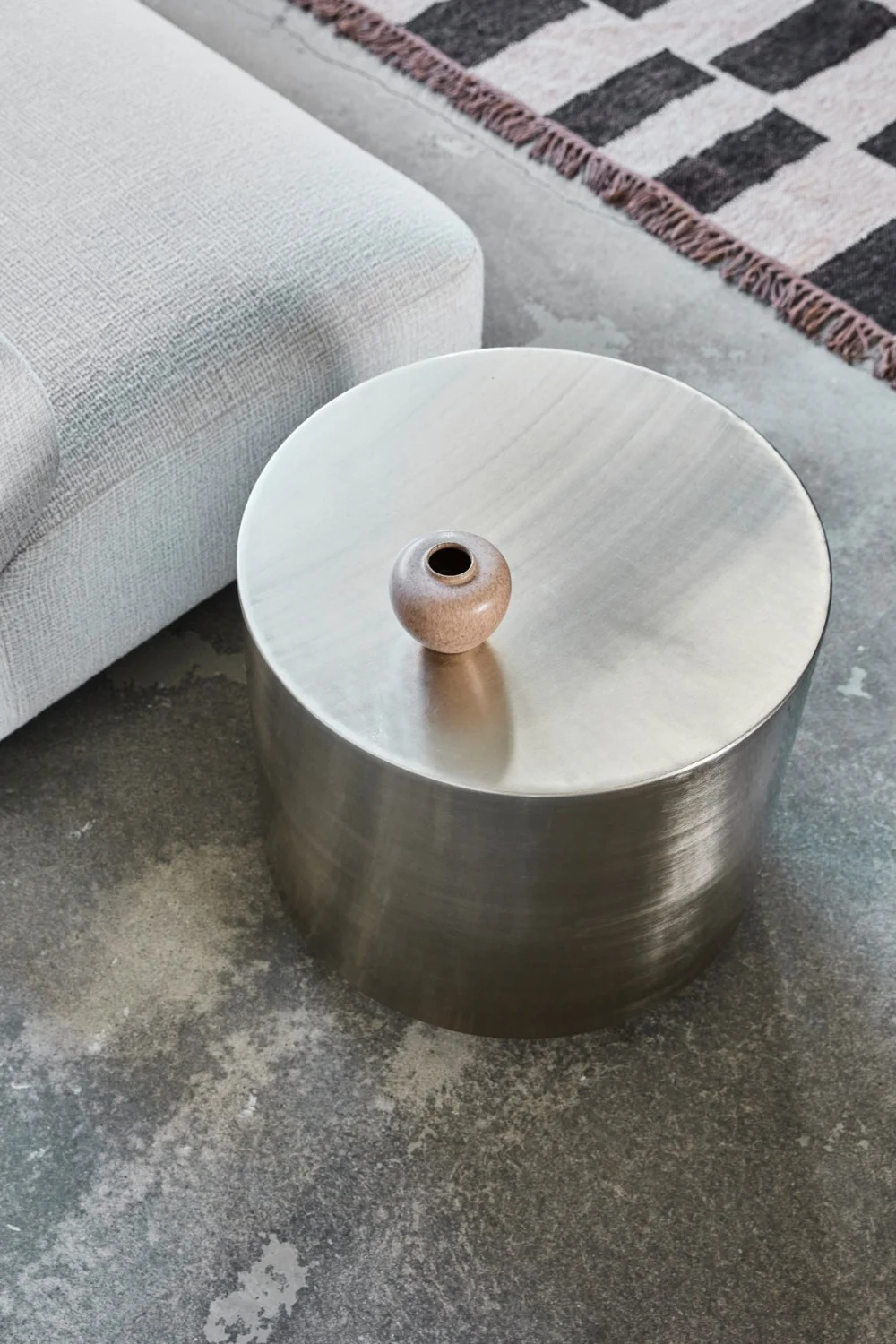 Une table d'appoint ronde en métal sur laquelle est posée une petite vasque en céramique, à côté d'un canapé gris clair et d'un tapis à motifs posé sur un sol en béton.