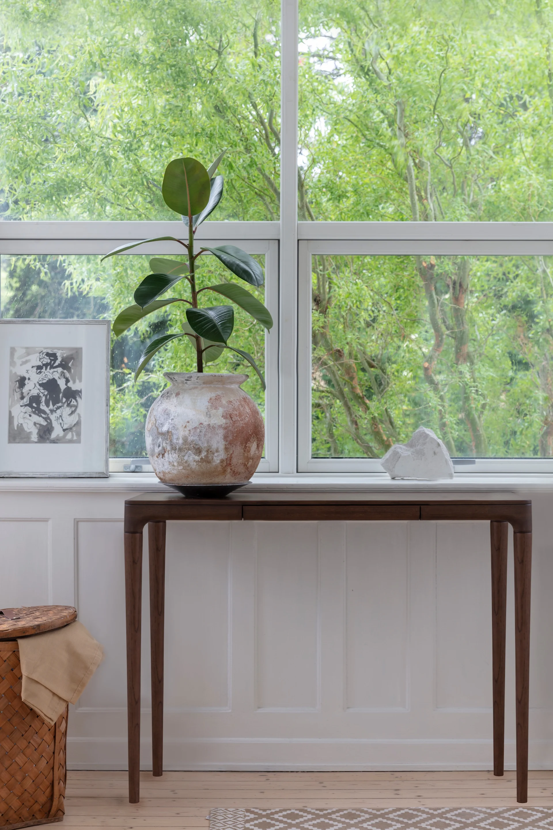 Une plante en caoutchouc dans un pot rustique sur une console près d'une fenêtre, avec une œuvre d'art encadrée et une sculpture blanche. Des arbres verts sont visibles à l'extérieur.