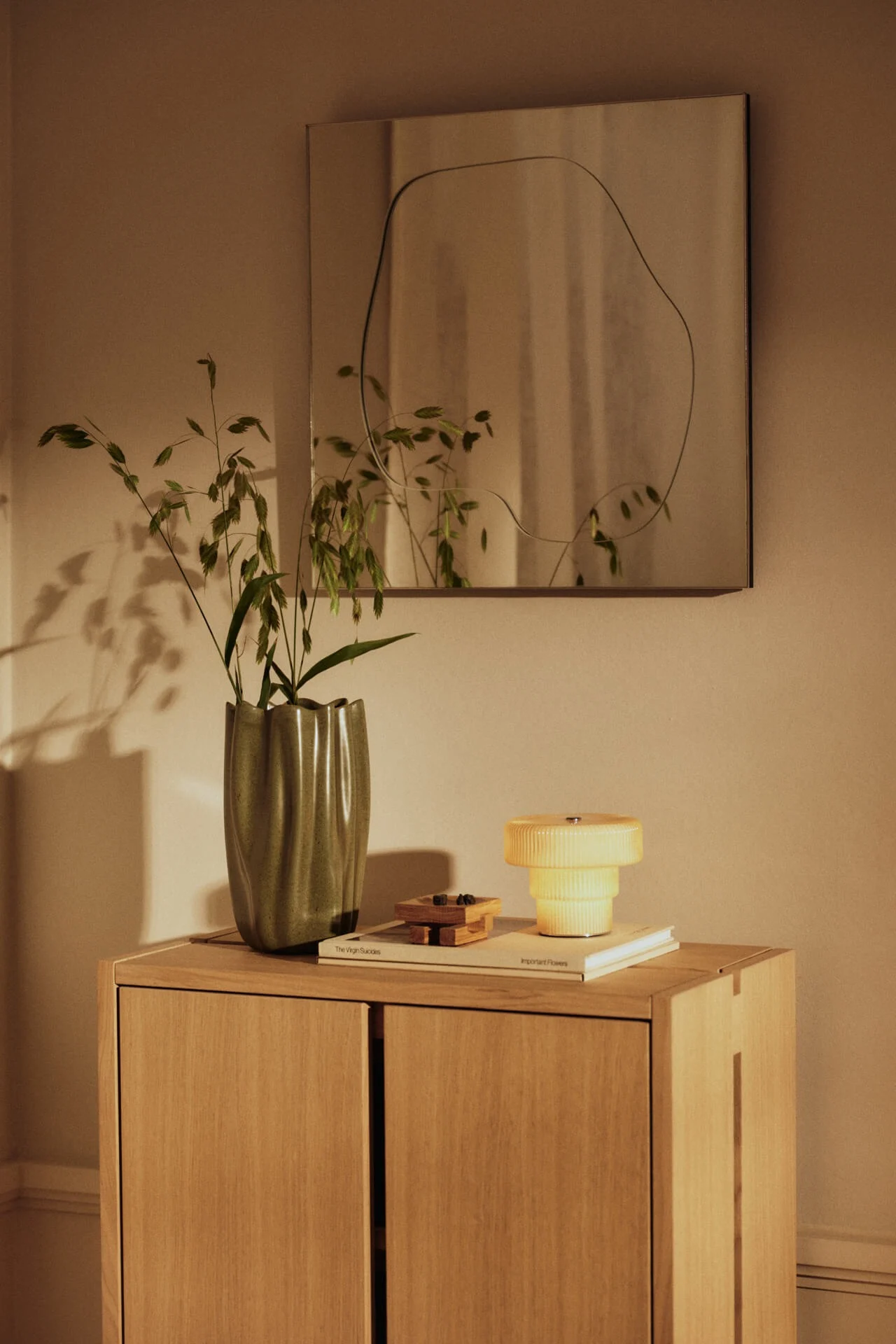 Un meuble en bois avec un vase vert ondulé de plantes, une lampe jaune étagée et un miroir au contour irrégulier sur le mur.