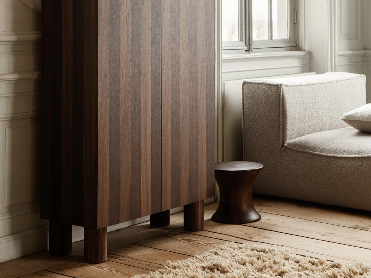 Pièce élégante avec armoire en bois foncé cannelée, sculpture de femme agenouillée, canapé modulaire clair et grande fenêtre.