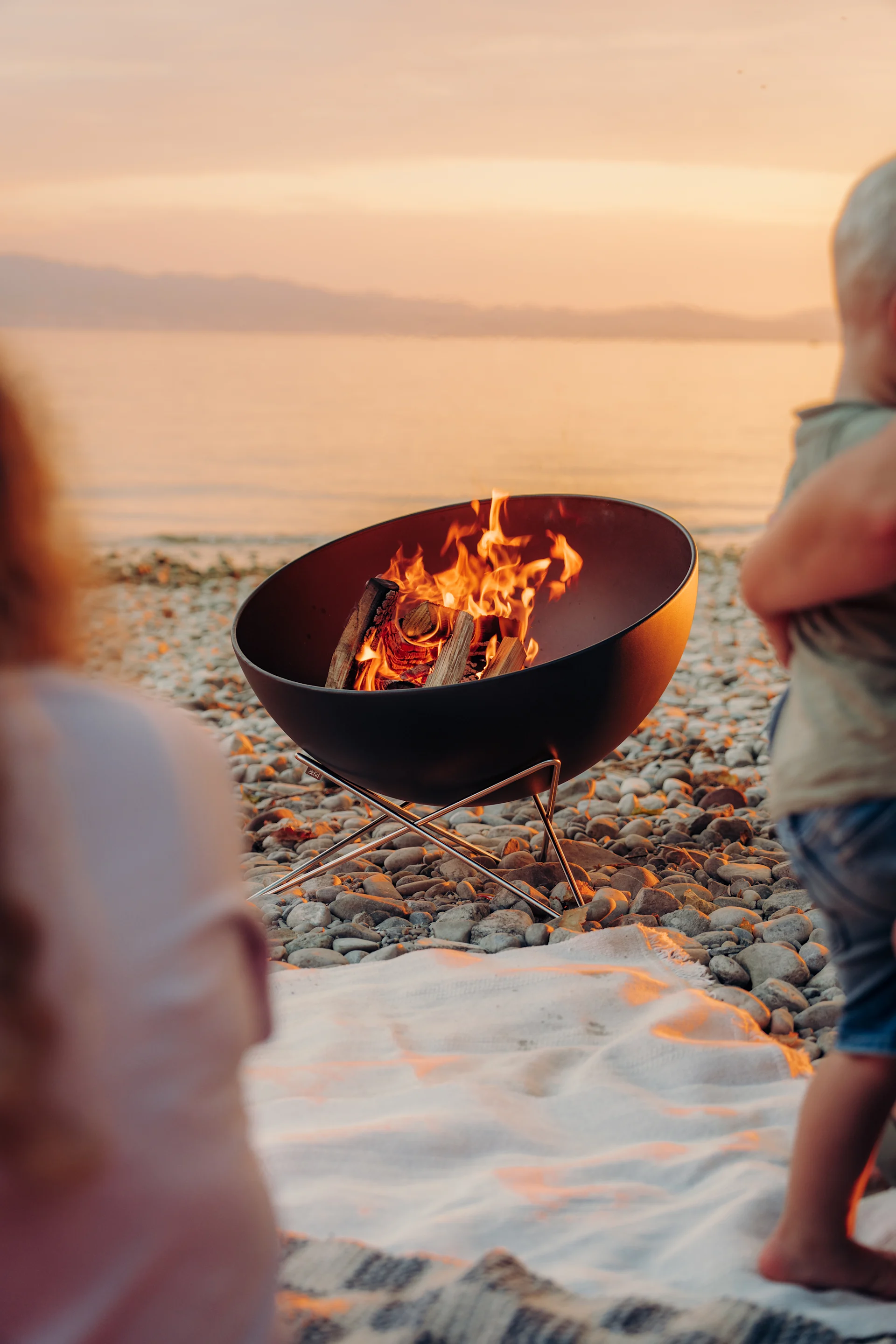 Un brasero aux flammes rougeoyantes sur une plage de galets au coucher du soleil, avec des silhouettes floues au premier plan.