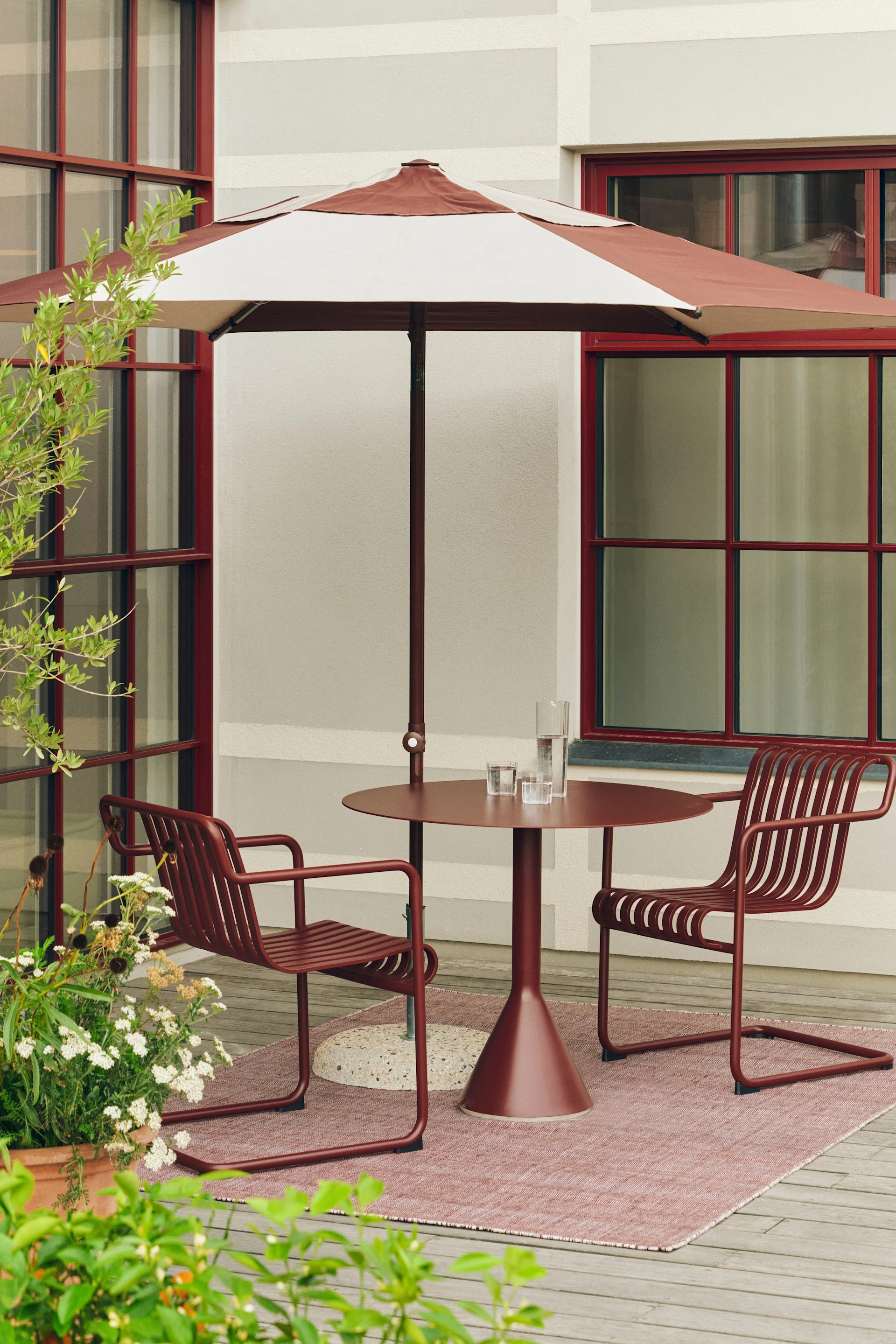 Terrasse extérieure avec parasol rayé marron-rouge, table ronde assortie et deux chaises à lattes sur tapis rose.