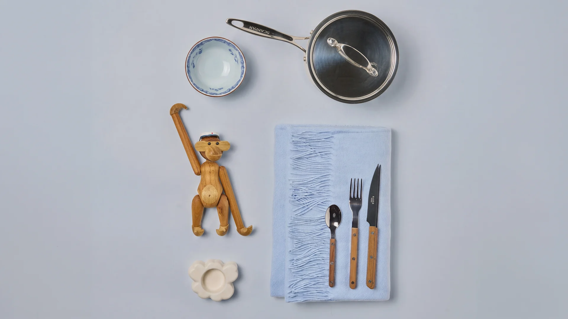 Casserole SCANPAN, bol bleu, singe en bois, bougeoir en forme de nuage et couverts sur un tissu bleu clair.