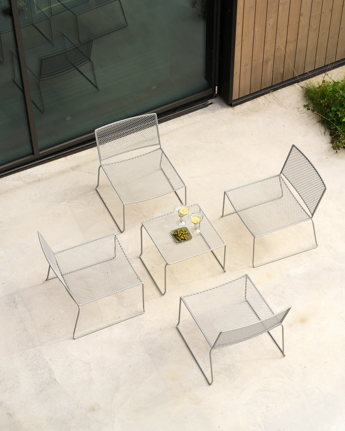 Salon extérieur gris clair en vue plongeante: quatre chaises grillagées, table avec boissons et olives sur patio.