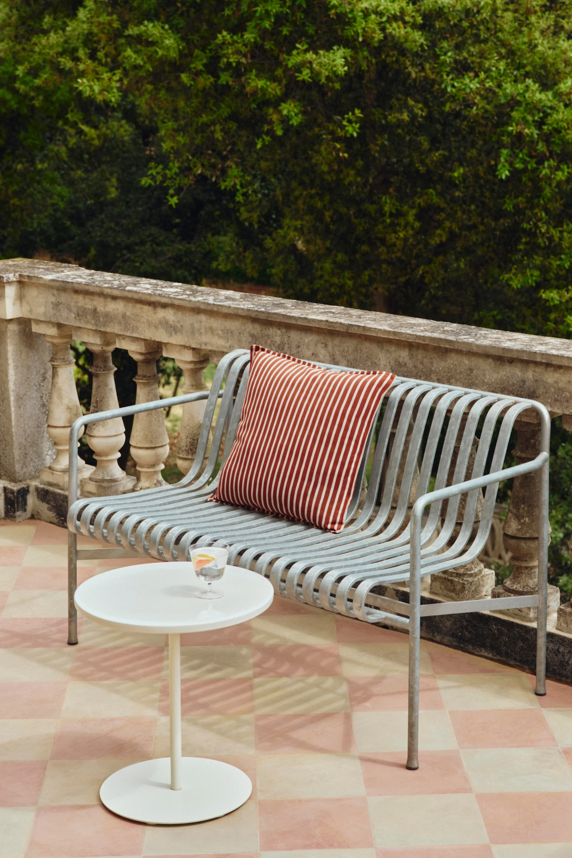 Un banc extérieur en métal gris clair avec un coussin rayé rouge et blanc, à côté d'une table d'appoint blanche avec une boisson sur un patio.