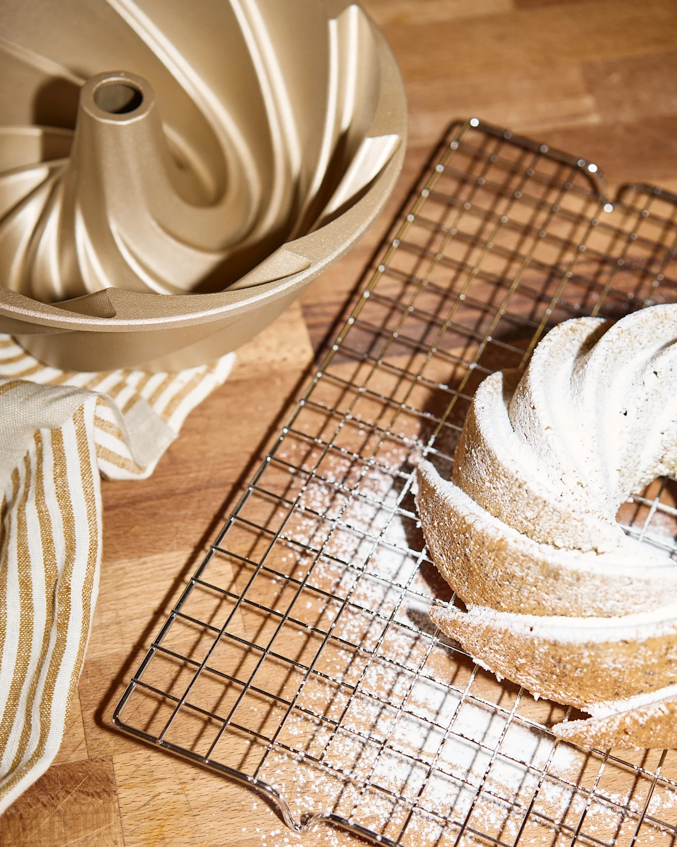 Un moule à bundt cake doré, une grille de refroidissement avec un bundt cake saupoudré de sucre glace et une serviette rayée sur une surface en bois.