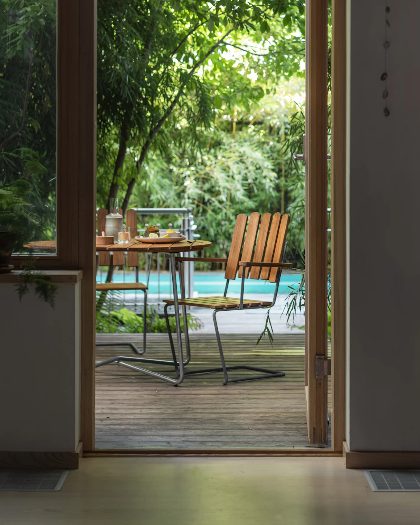 Vue à travers une porte ouverte sur une terrasse extérieure avec une table en bois, des chaises et une piscine entourée de plantes vertes.