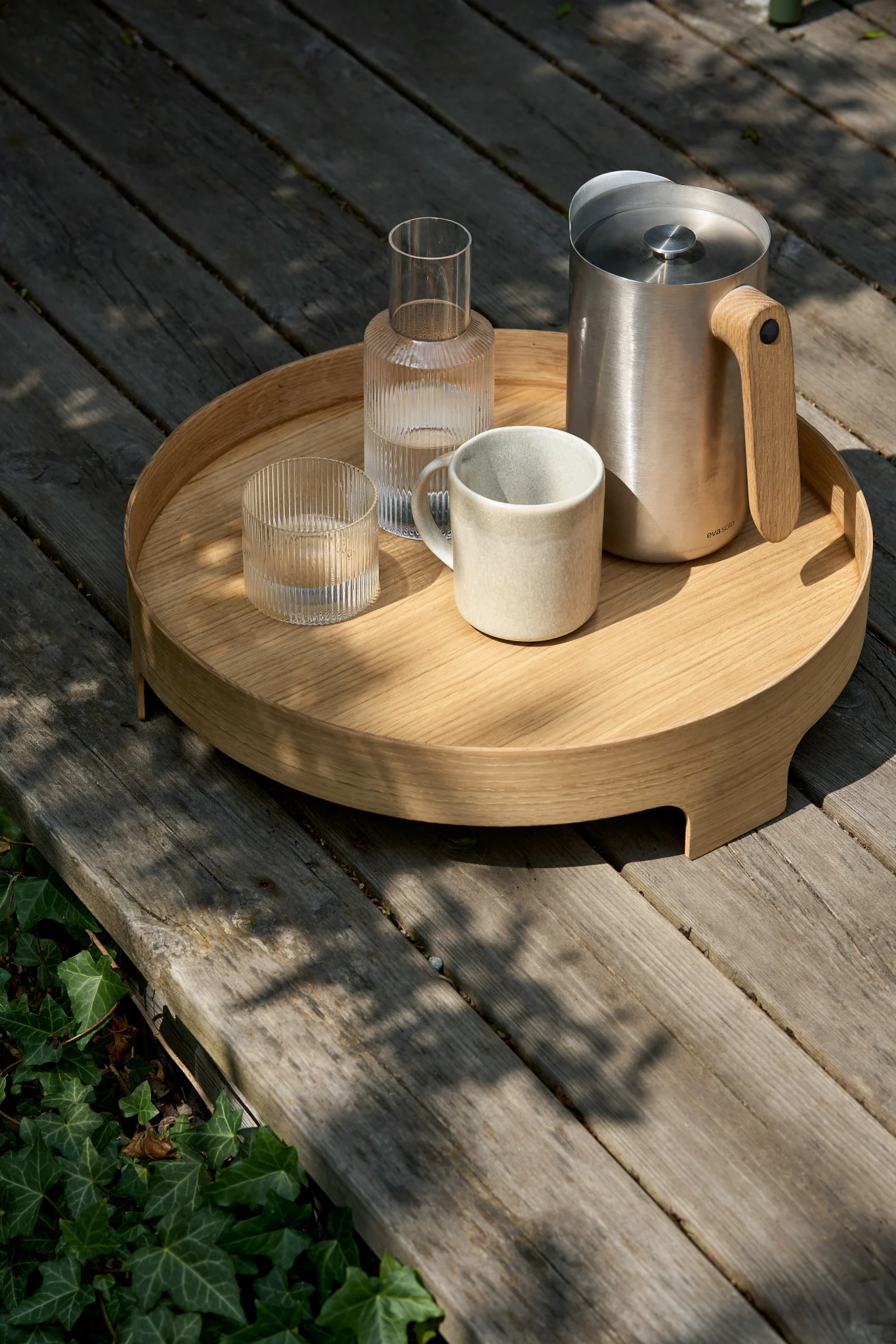 Sur une table en bois, un plateau rond accueille un pichet en acier inoxydable muni d'une poignée en bois, une carafe en verre strié, un verre strié et une tasse en céramique.
