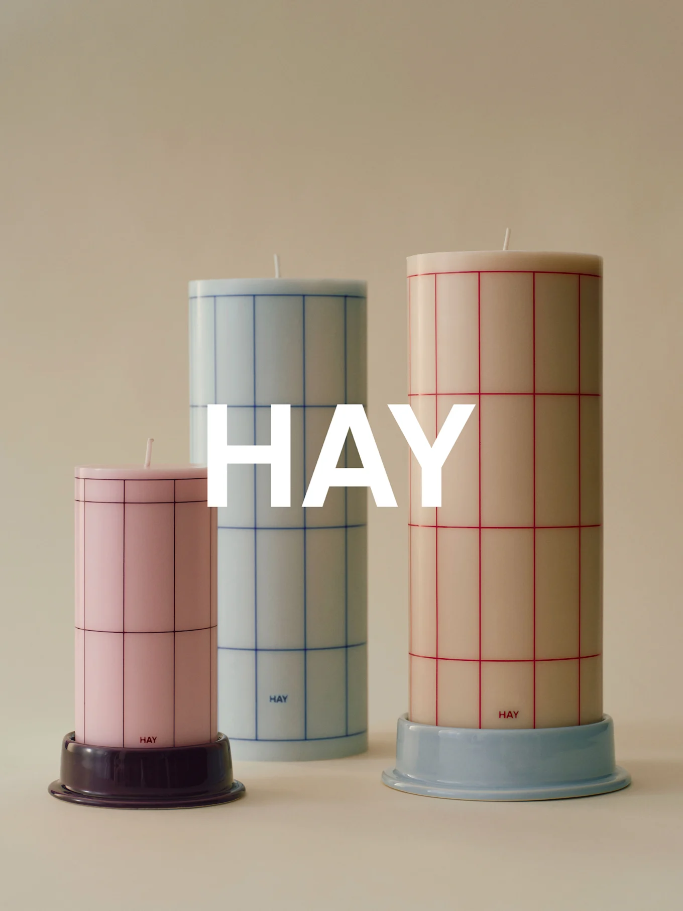 Trois bougies cylindriques à motif quadrillé de différentes hauteurs: rose, bleu clair et crème, sur socles.