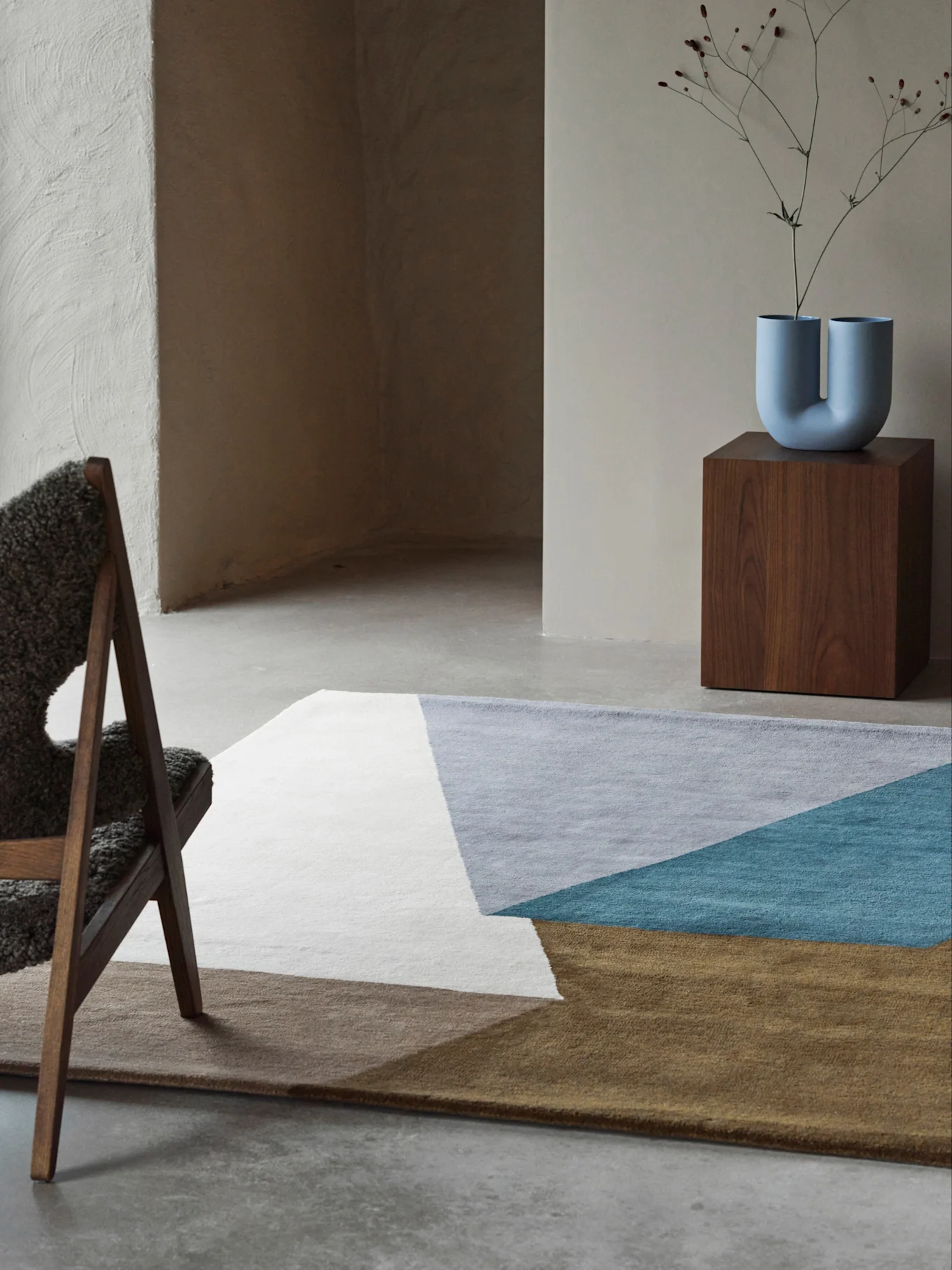 Pièce moderne avec un tapis géométrique blanc, gris, bleu sarcelle et marron, un vase bleu en forme de U sur un piédestal en bois et une chaise moelleuse.