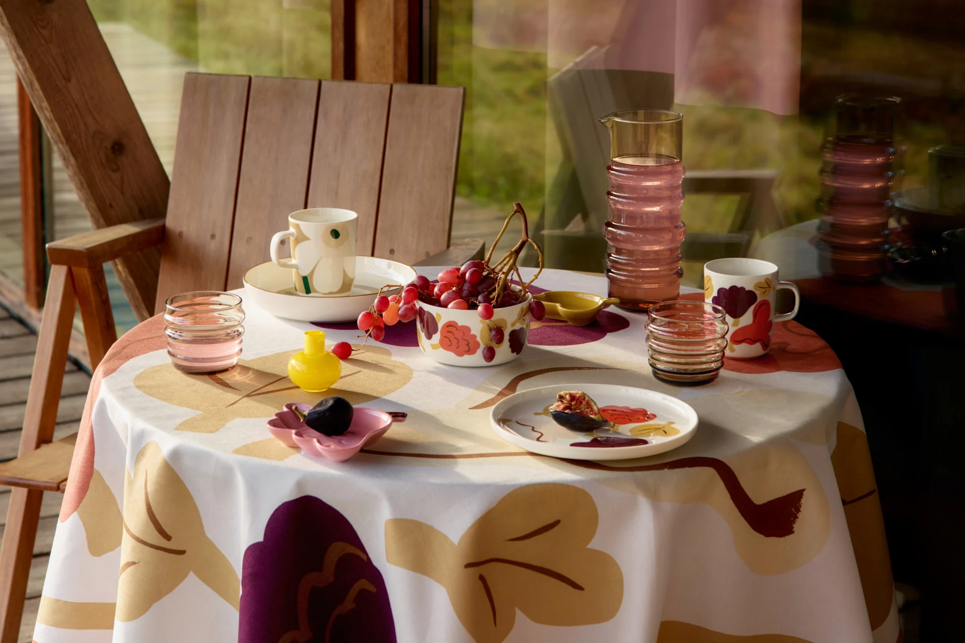 Une table dressée avec une nappe à motifs, des tasses, des bols de raisin, une figue sur une assiette, une carafe et des verres, à côté d'une chaise en bois.