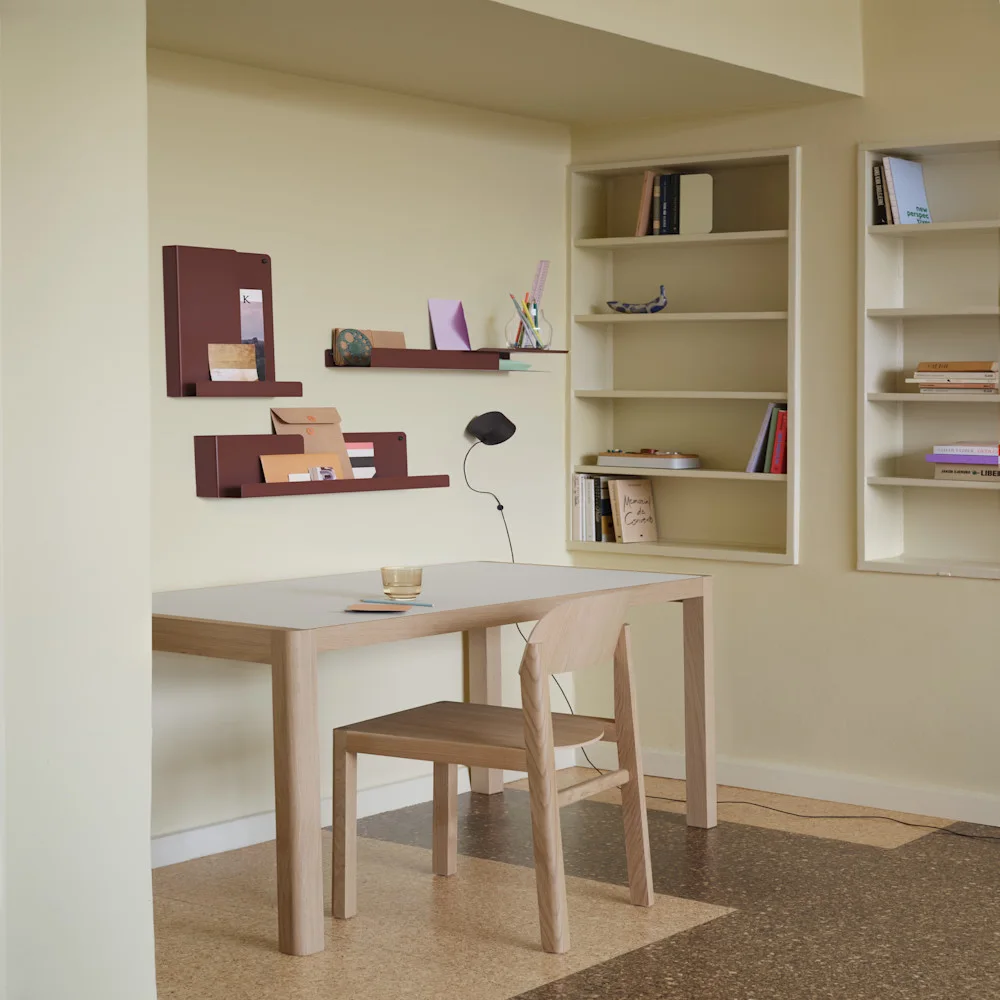 Un bureau minimaliste avec une table en bois clair, une chaise et trois étagères murales brun rougeâtre. Des bibliothèques intégrées sont à droite.