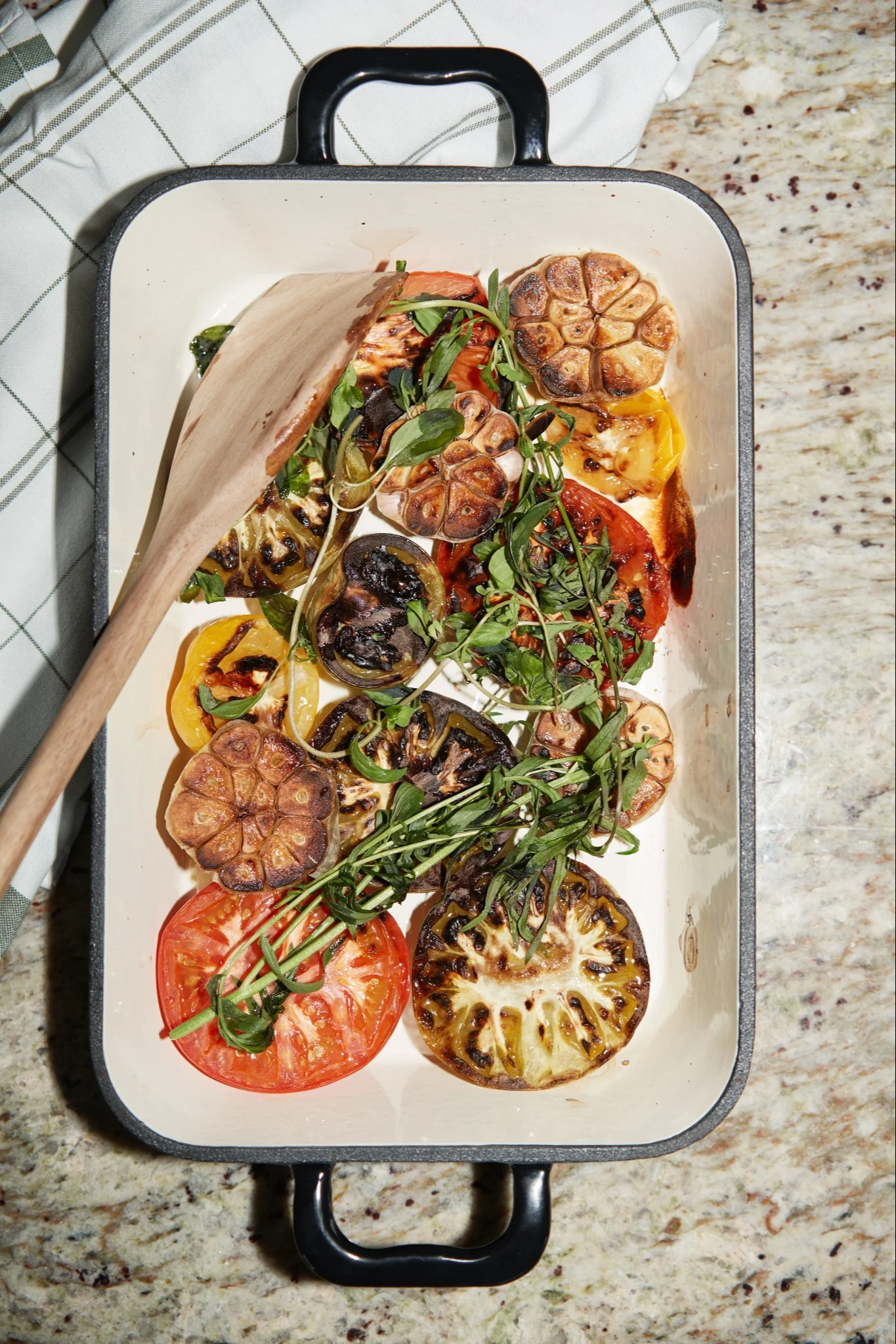 Un plat à four blanc rempli de tomates grillées, d'aubergines, d'ail rôti et d'herbes aromatiques fraîches, avec une cuillère en bois.