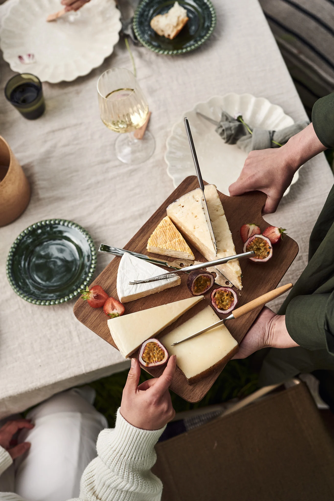 Des mains disposent sur une table dressée un plateau à fromage en bois garni de différents fromages, de fraises coupées en deux et de fruits de la passion.