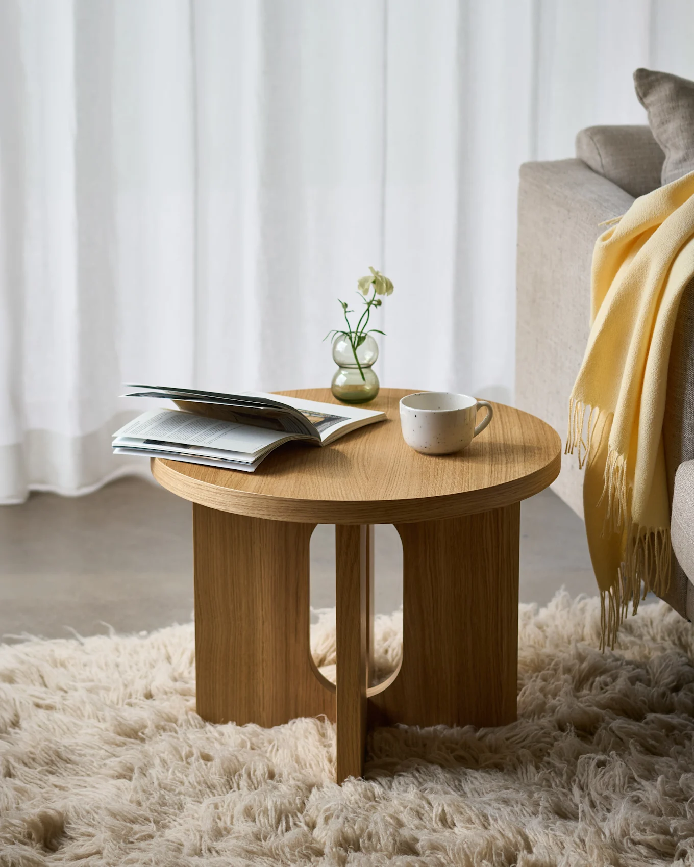 Table d'appoint ronde en bois sur un tapis moelleux, avec un magazine ouvert, une fleur blanche dans un vase et une tasse en céramique, à côté d'un canapé.