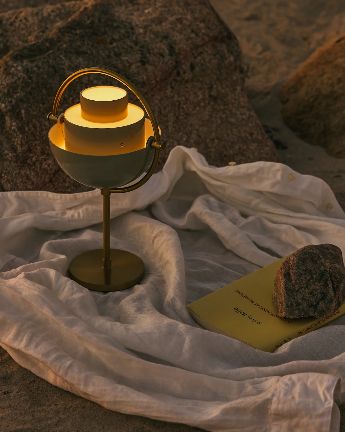 Une lampe de table moderne blanche et en laiton avec une lumière chaude, un livre jaune et une roche sur un tissu blanc sur une surface rocheuse.