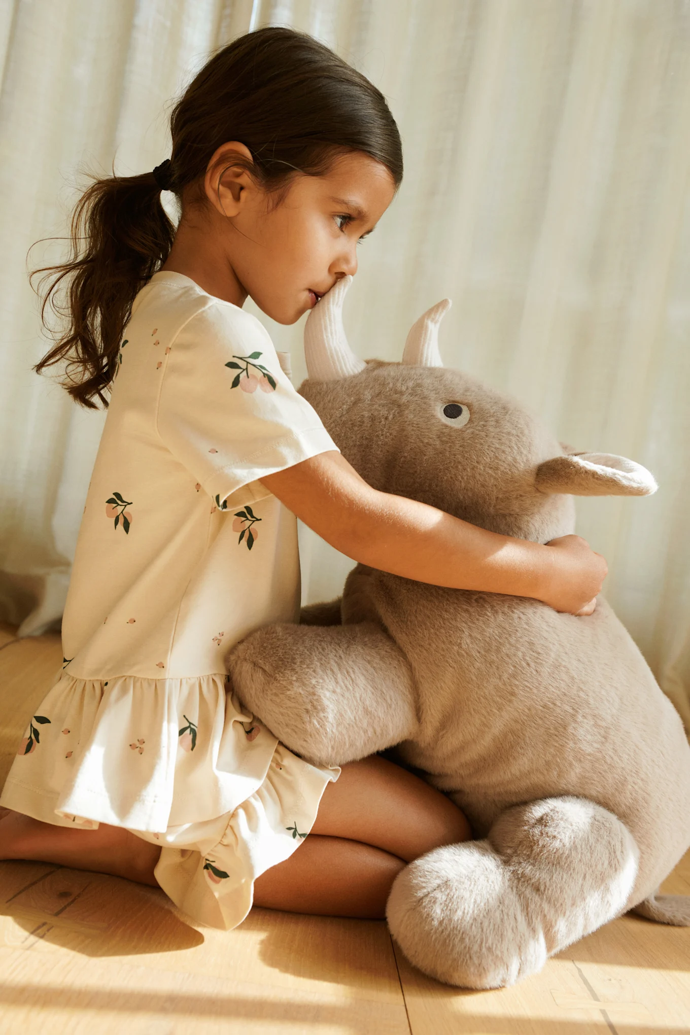 Jeune fille en robe à motifs agenouillée et serrant dans ses bras une grande peluche à cornes.
