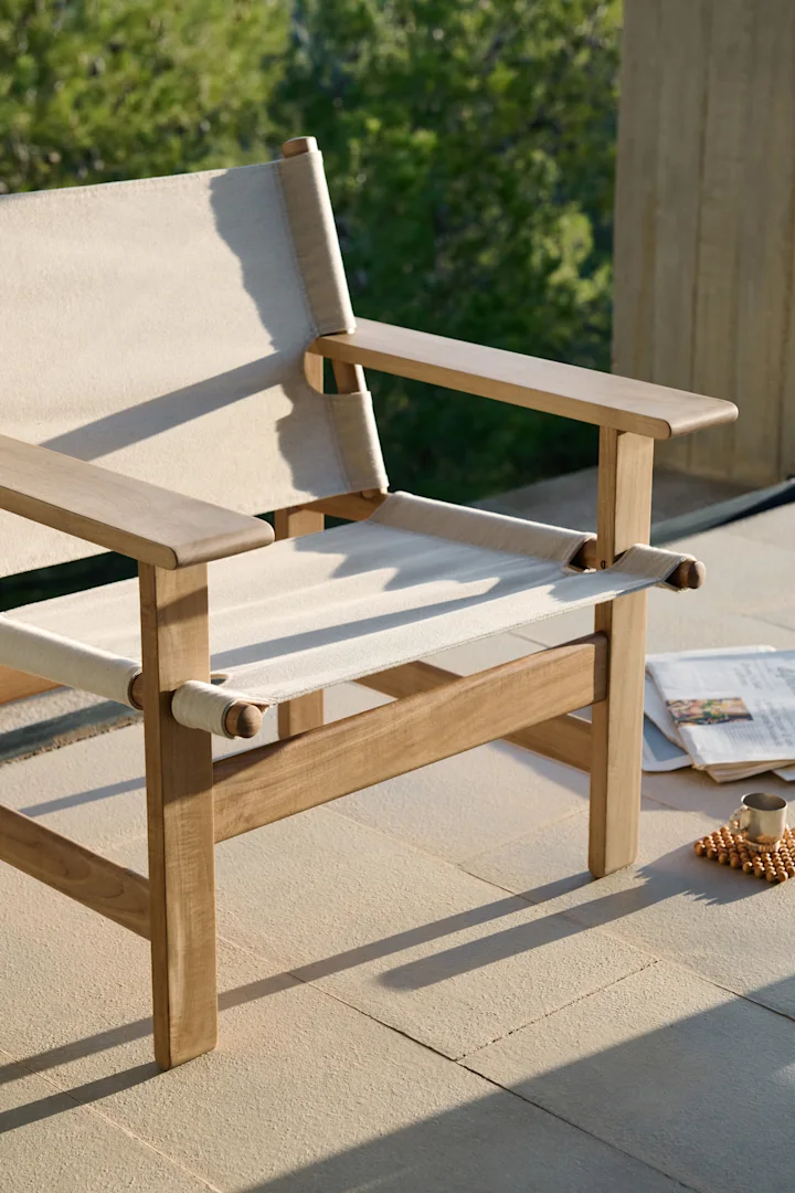 Chaise d'extérieur en bois clair avec toile crème sur une terrasse, avec un journal et une tasse à côté.