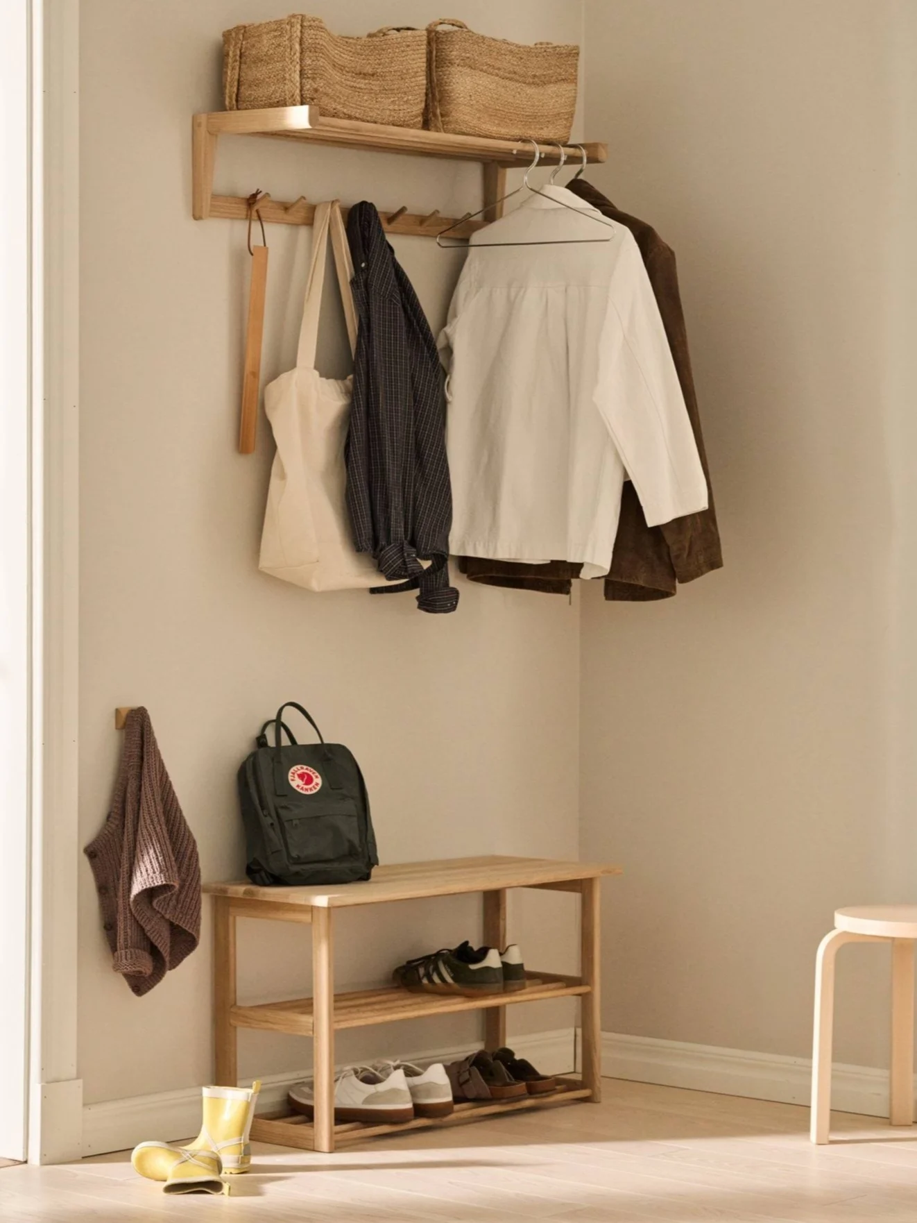 Un porte-manteau en bois avec des paniers tressés, des vêtements suspendus, un sac à dos vert, un meuble à chaussures avec baskets et sandales, et des bottes de pluie jaunes dans une entrée.