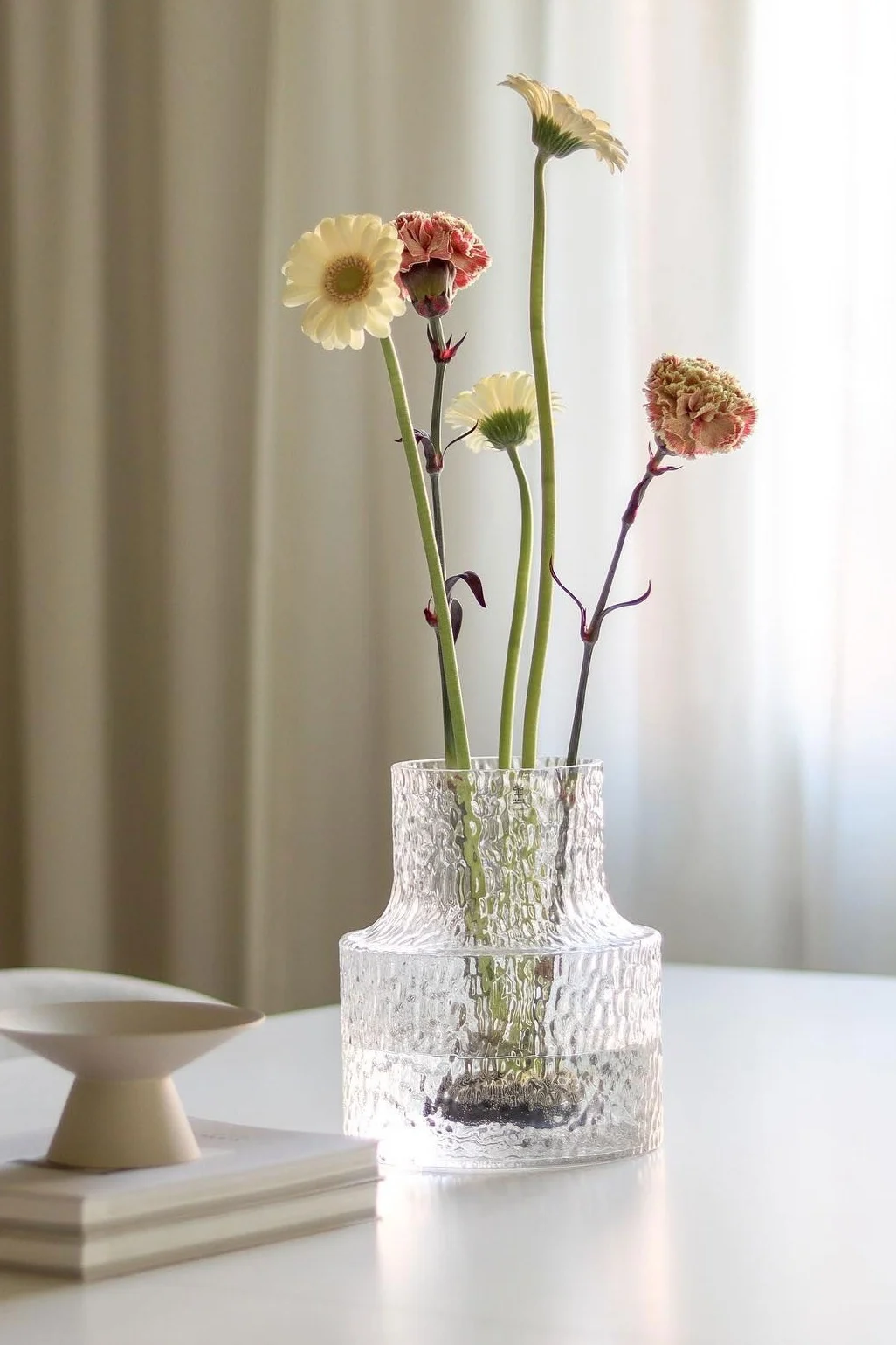Un vase en verre transparent texturé contient des gerberas jaunes pâles et des œillets rose-rouge sur une table.