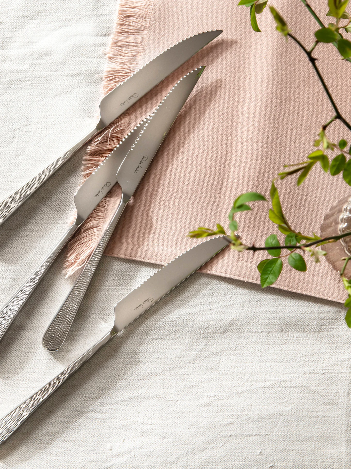 Quatre couteaux à steak en acier inoxydable avec lames dentelées et manches texturés sur une nappe en lin avec une serviette rose.