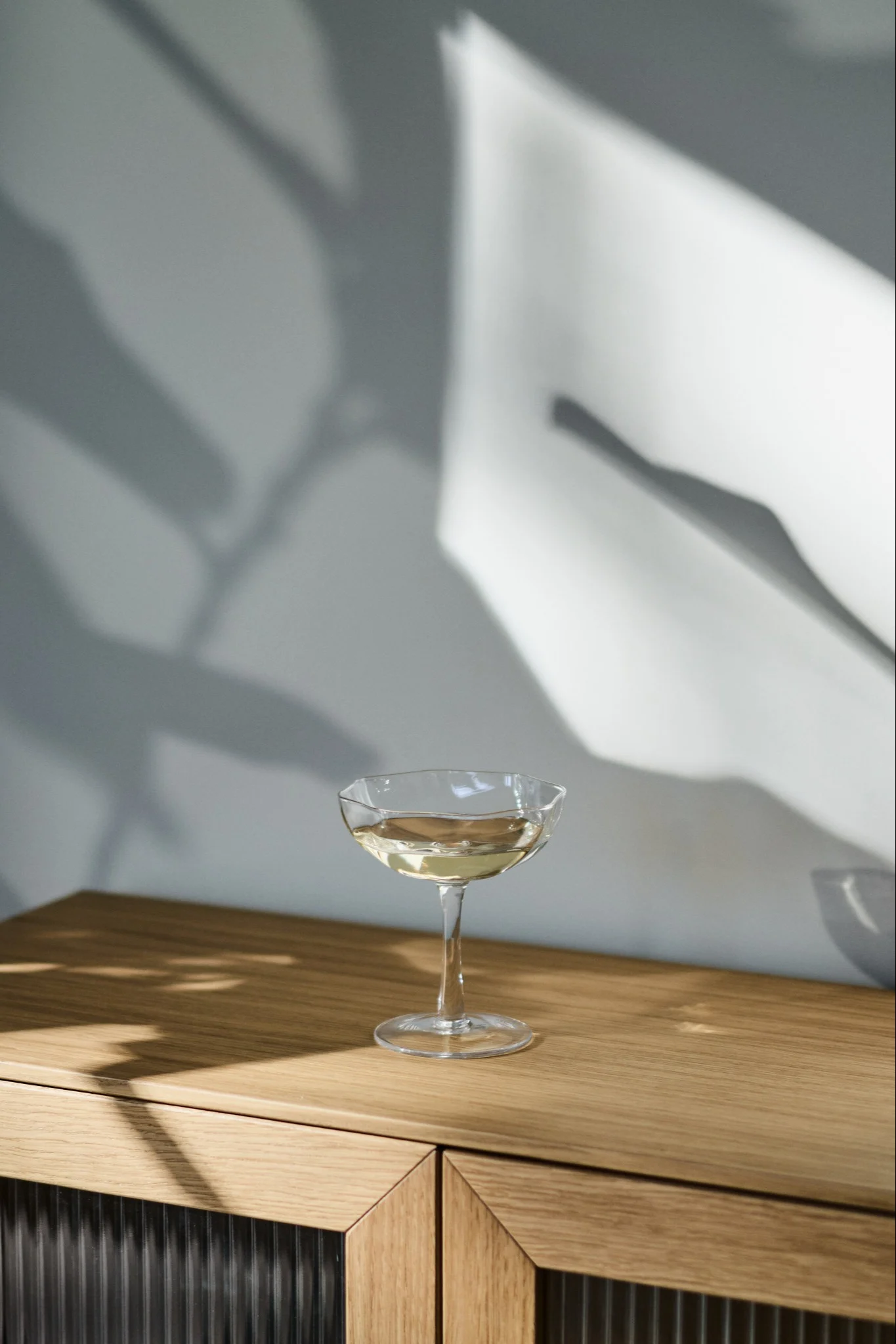 Une coupe avec une boisson claire sur une armoire en bois, avec des ombres abstraites sur le mur.