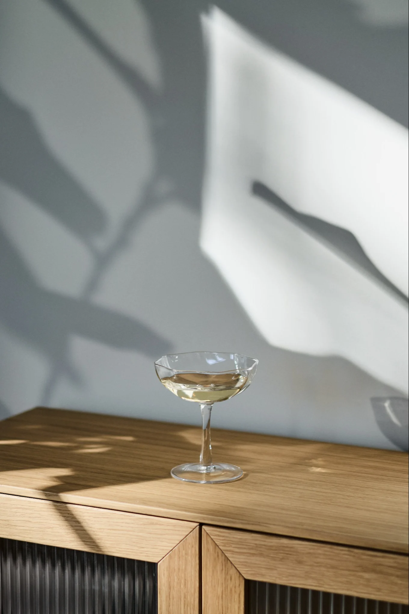 Une coupe avec une boisson claire sur une armoire en bois, avec des ombres abstraites sur le mur.