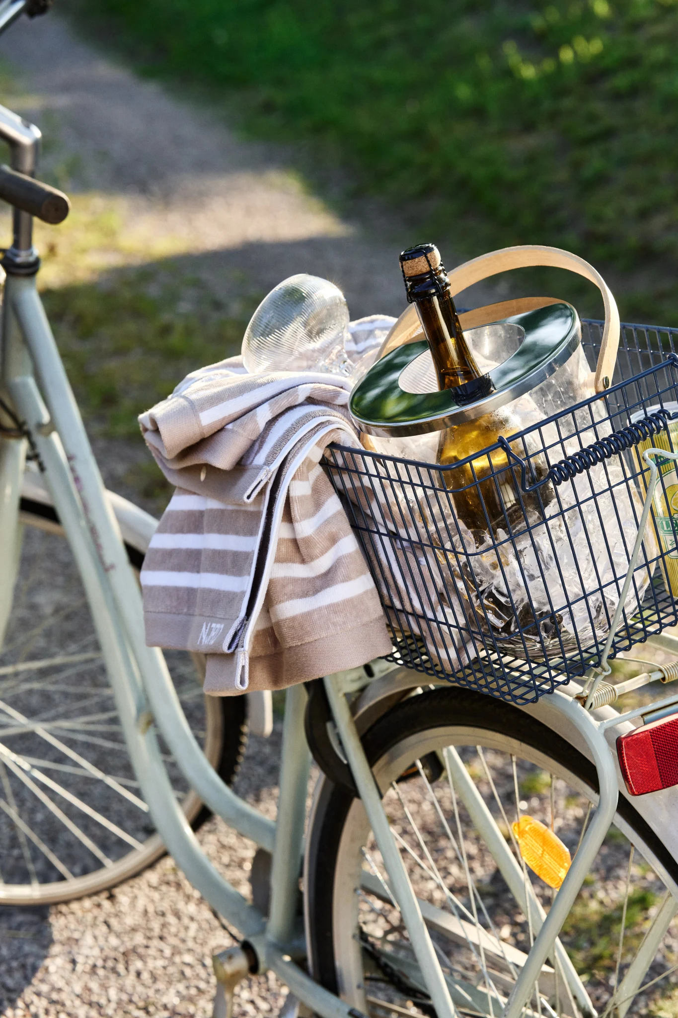 Un panier de vélo contient un seau à glace avec une bouteille réfrigérante et une serviette rayée, par une journée ensoleillée.